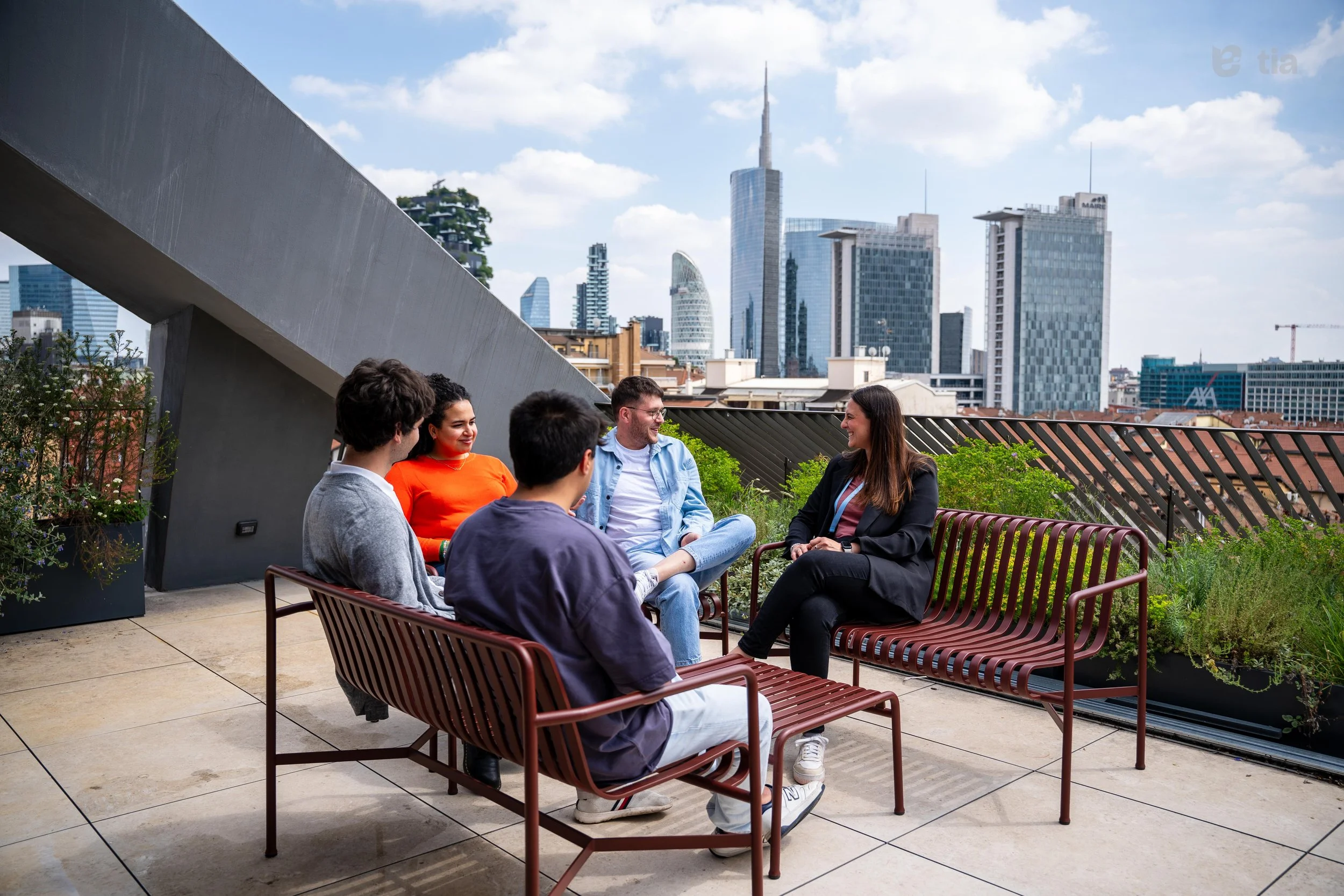Gruppo di cinque persone che siedono su sedie di metallo rosso su un tetto di un edificio con vista sulla città di notte e alcuni grattacieli al sole. Parlando e sorridendo insieme.