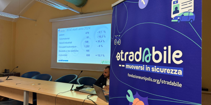 Sala con tavolo e sedie, proiettore con presentatione sul muro, persona seduta al tavolo, banner con testo 'stradability - muoversi in sicurezza'.