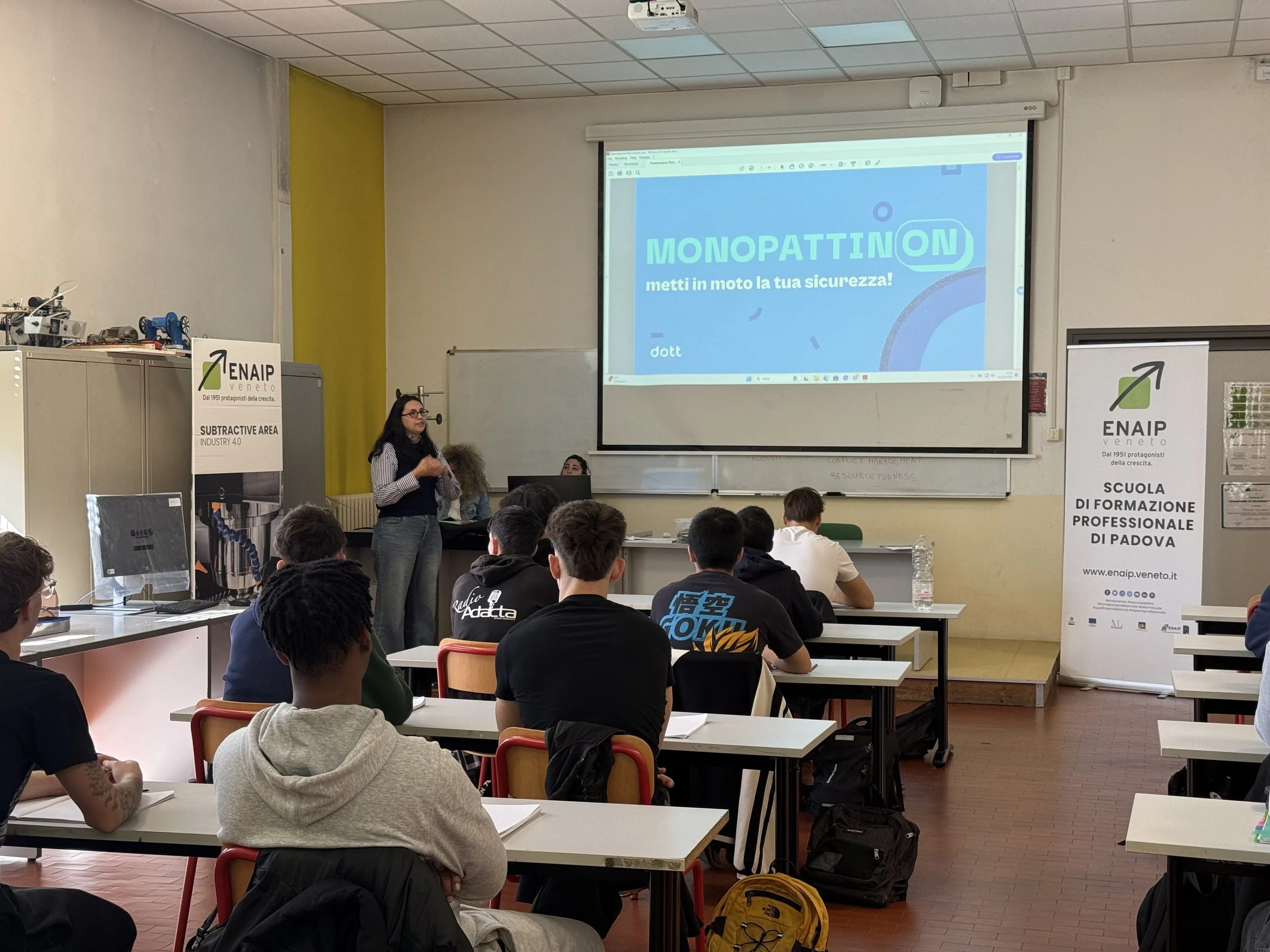 Una lezione in aula con studenti seduti ai banchi e una docente in piedi di fronte a una presentazione proiettata su uno schermo. Vi sono anche due banner laterali con logo e testo promozionale.