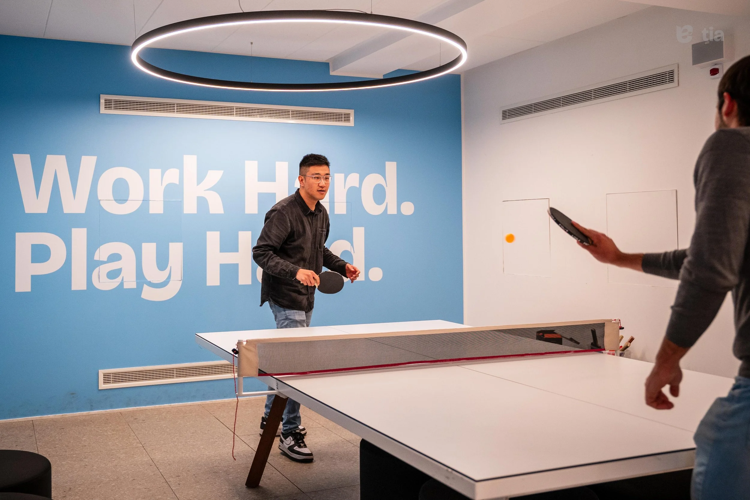 Due persone che giocano a ping pong in una stanza con pareti bianca e blu, con motto "Work Hard. Play Hard." scritto sul muro. Uno sta colpendo la palla arancione, l'altro si sta preparando a rispondere.