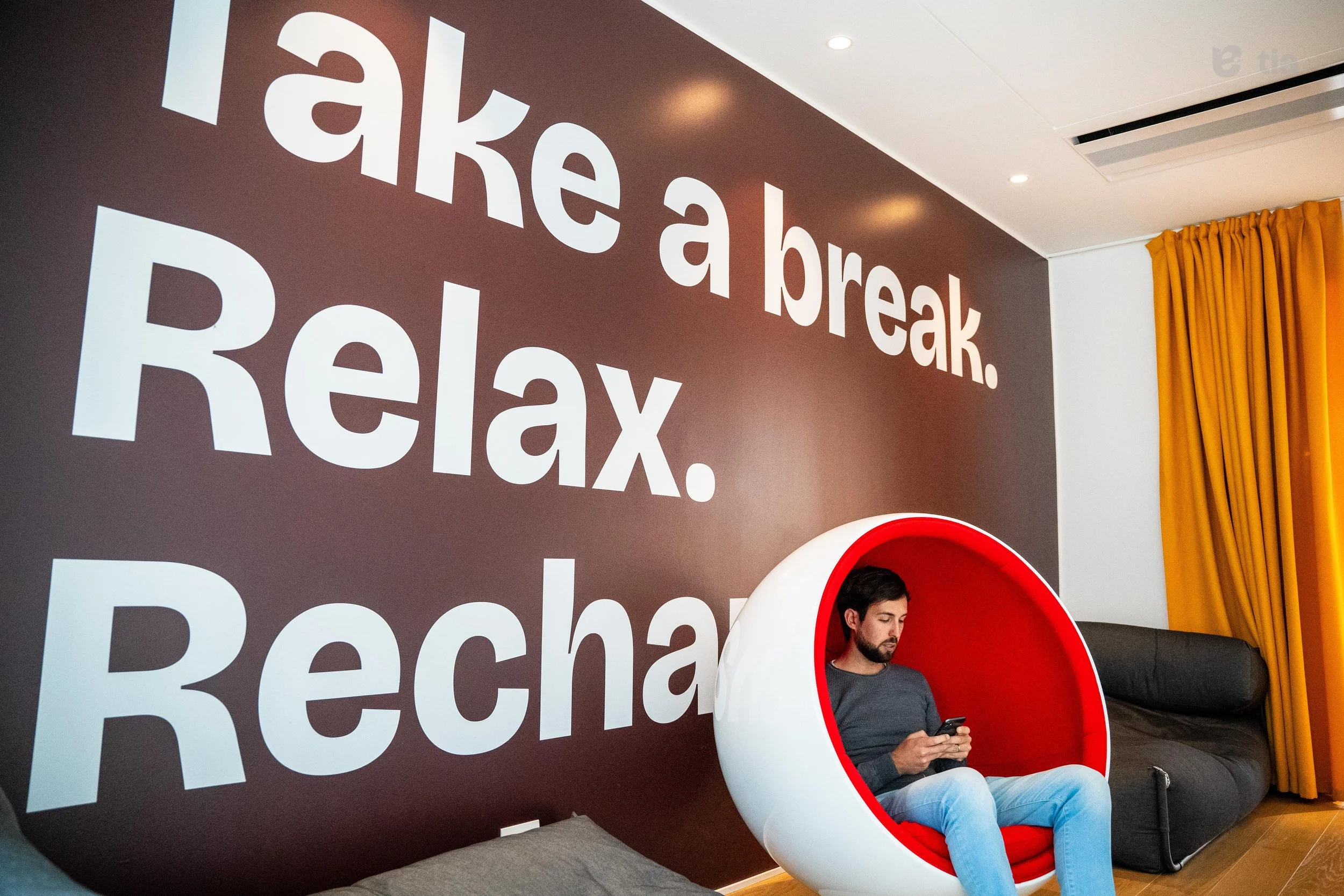 Uomo seduto in una sedia a forma di metà sfera rossa e bianca, che guarda lo smartphone, in una stanza con parete marrone con scritta bianca 'take a break. Relax. Recharge.' e tende gialle.