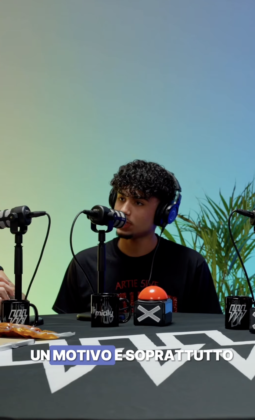 Un giovane uomo con capelli ricci scuri indossa cuffie, seduto di fronte a tre microfoni, con una tazza nera e un pulsante rosso sul tavolo.