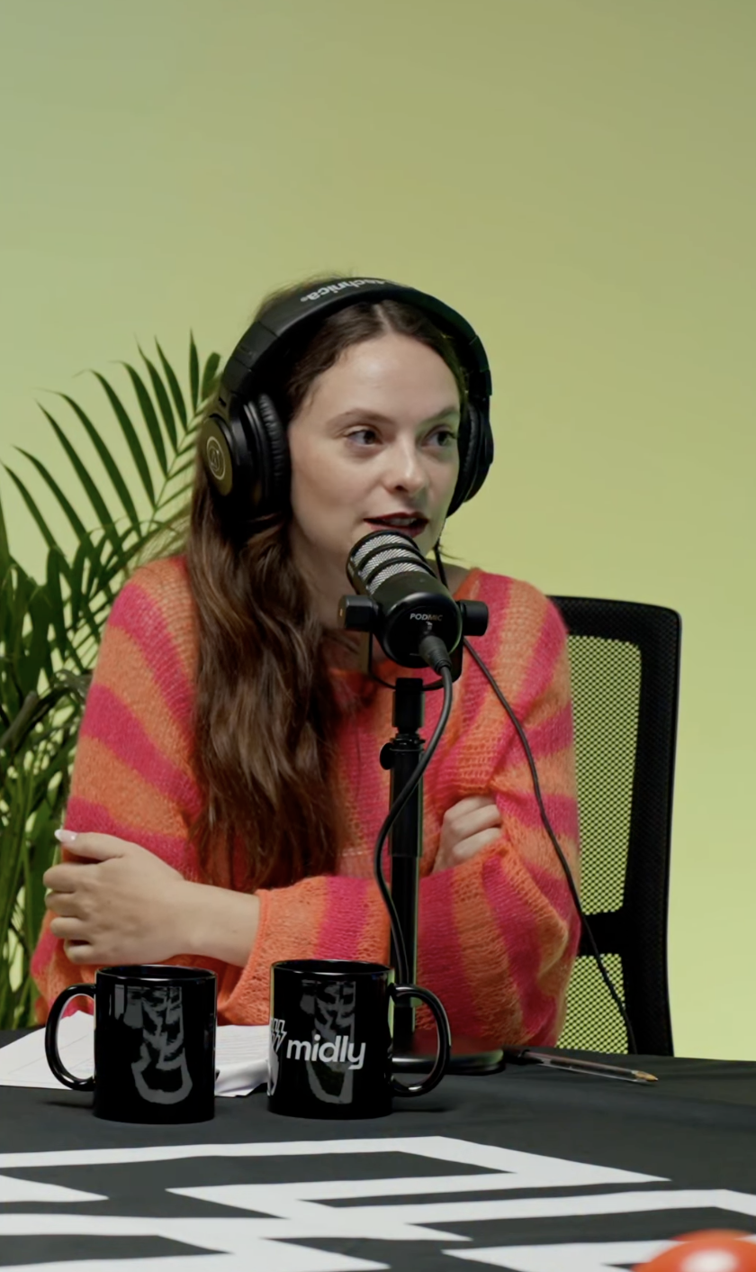 Donna con lunghi capelli castani e maglione a righe rosa e arancioni ascolta e parla in un podcast, con cuffie e microfono, seduta a un tavolo con due tazze nere con il logo 'midly', sullo sfondo parete verde chiaro e pianta