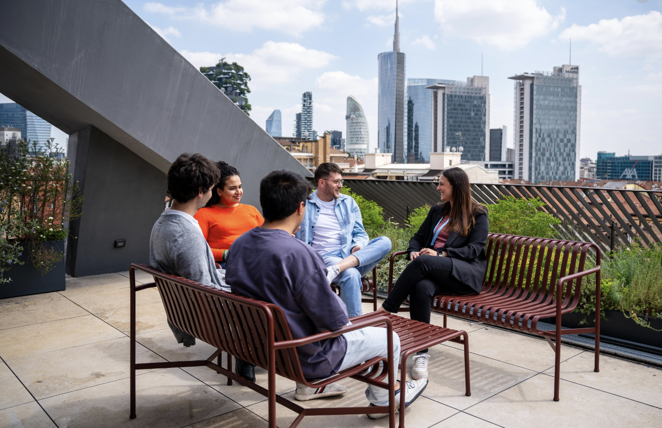 Cinque persone sedute su panchine di metallo su un terrazzo con vista sul skyline di una città, discutendo e sorridendo in un giorno soleggiato.