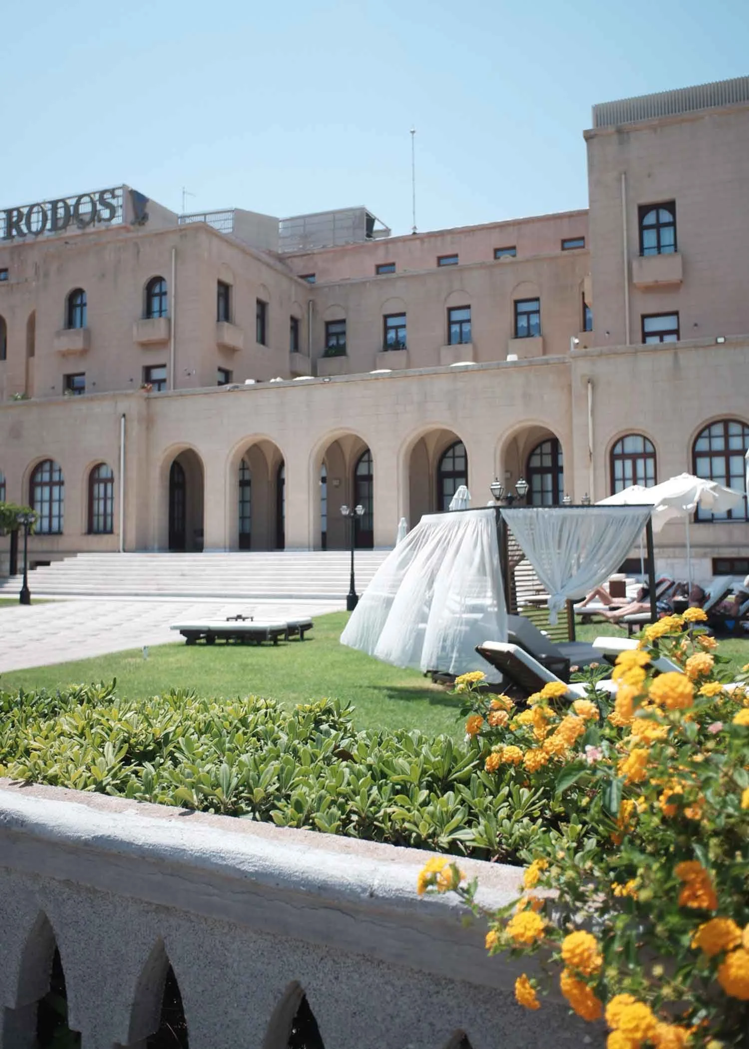 A hotel courtyard with a large stone building, steps leading up to the entrance, sun loungers with white canopies, and yellow flowers in the foreground.
