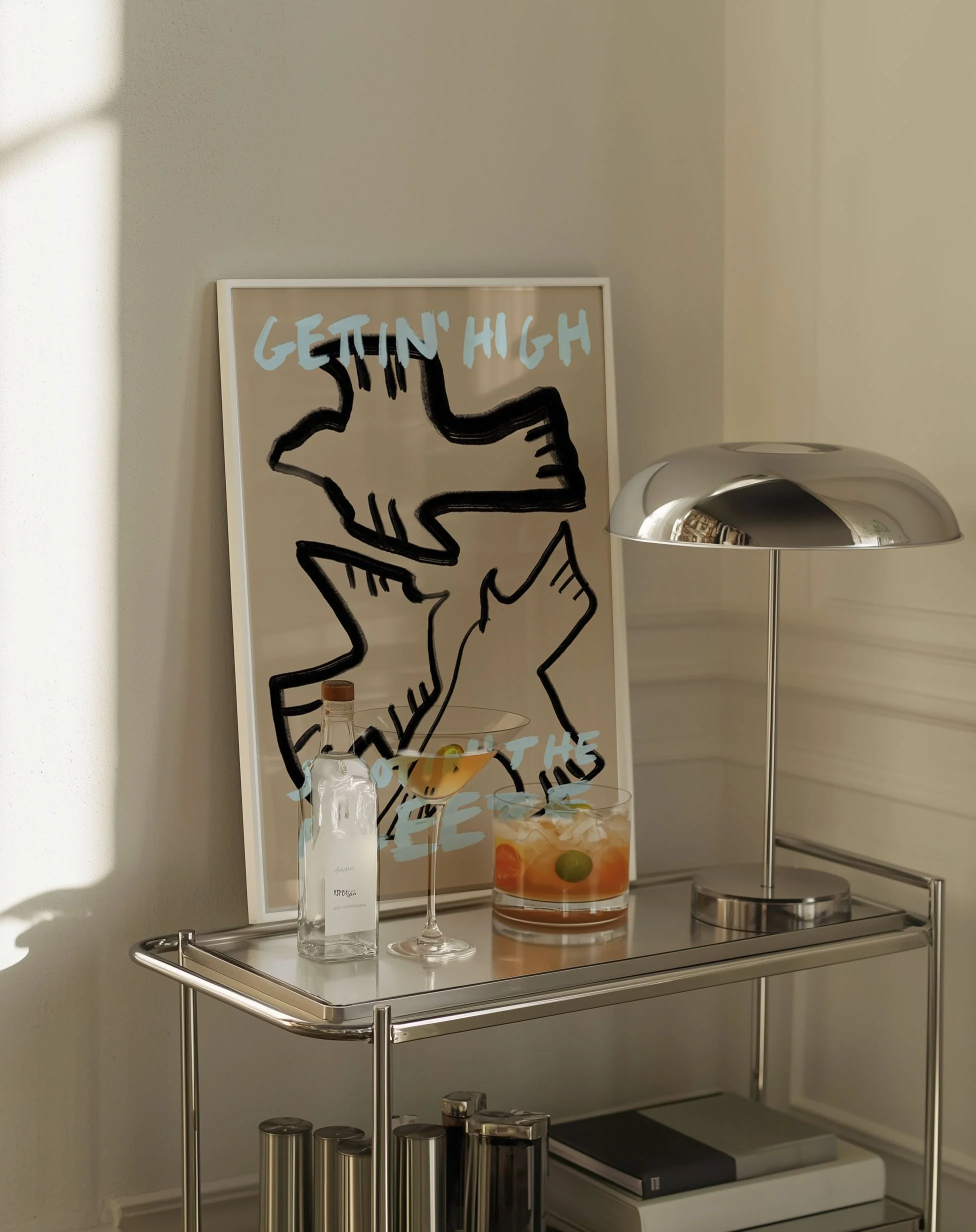 Minimalist typography poster featuring hand-drawn birds in black and beige with pastel blue lettering. poster in frame on a trendy barcart next to cocktails and chrome table lamp