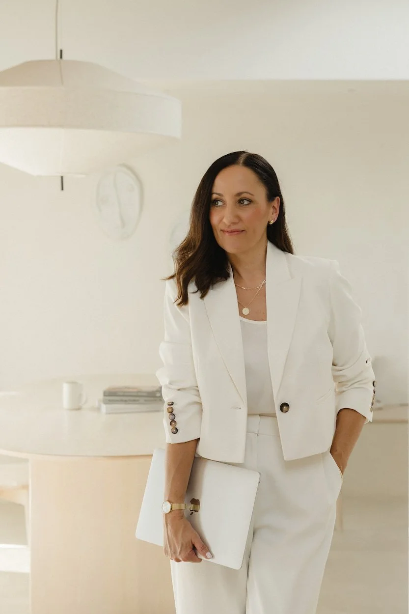 woman standing wearing a cream trouser suit holding an apple laptop