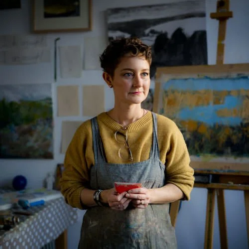 female artist wearing a muscard coloured jumper in from of canvas looking off to the side