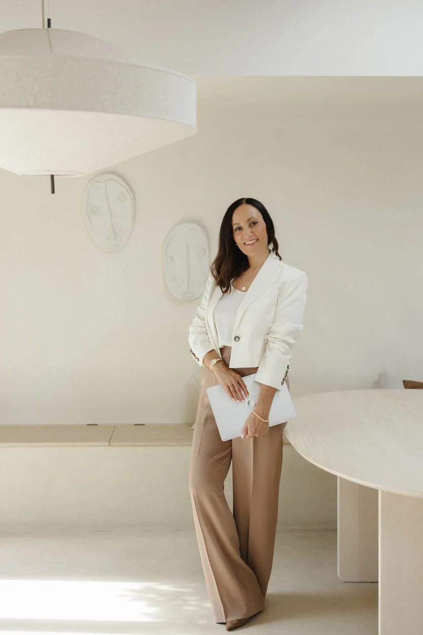 woman wearing a cream jacket and tan trousers holding a laptop on a stylish cream room