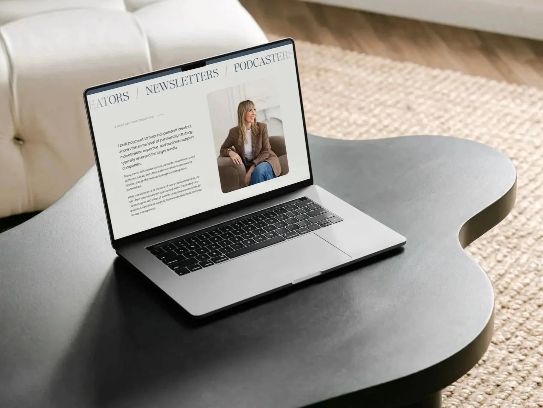 open laptop resting on a curved black table showing a neutral website featuring a woman in a tan jacket