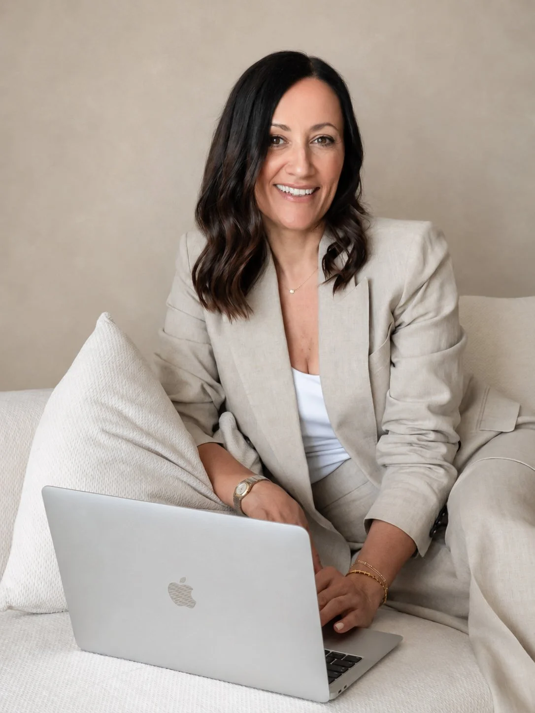 woman wearing a linen trouser suit seated on a cream sofa, working on a laptop