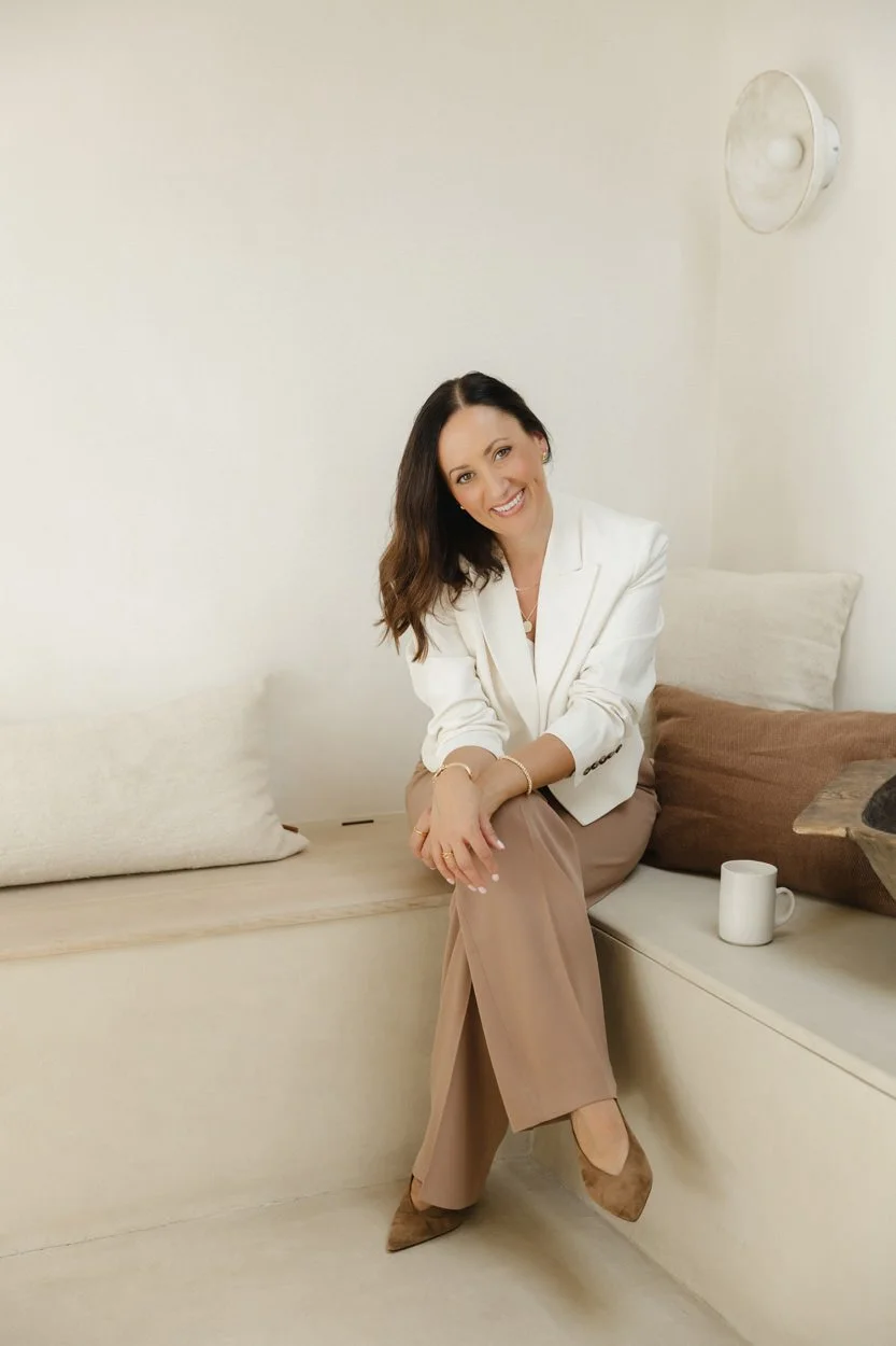 brunette woman wearing a white jacket and tan trousers sitting on a cream indoor bench