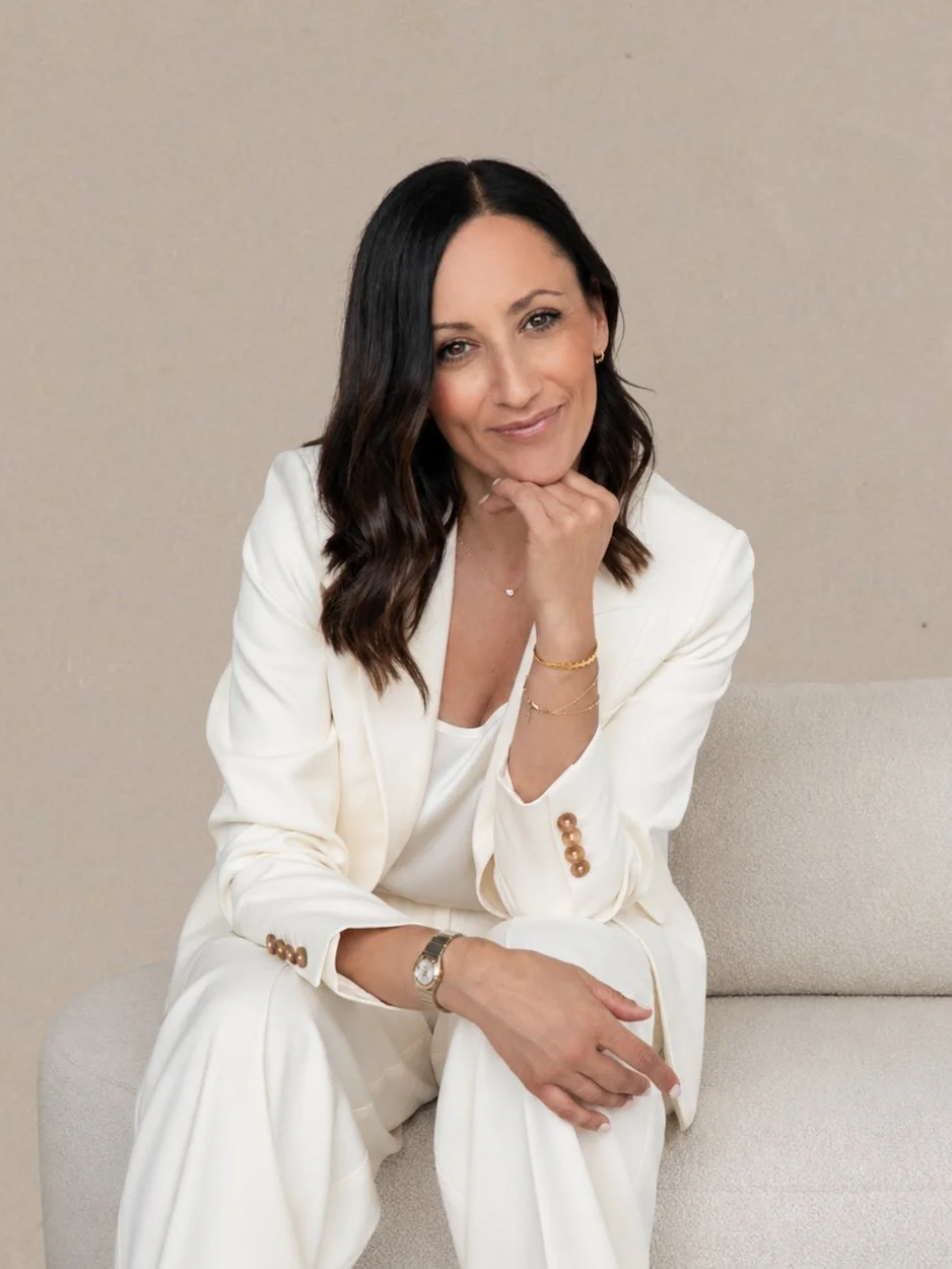brunette woman wearing a white suit sitting on a cream sofa