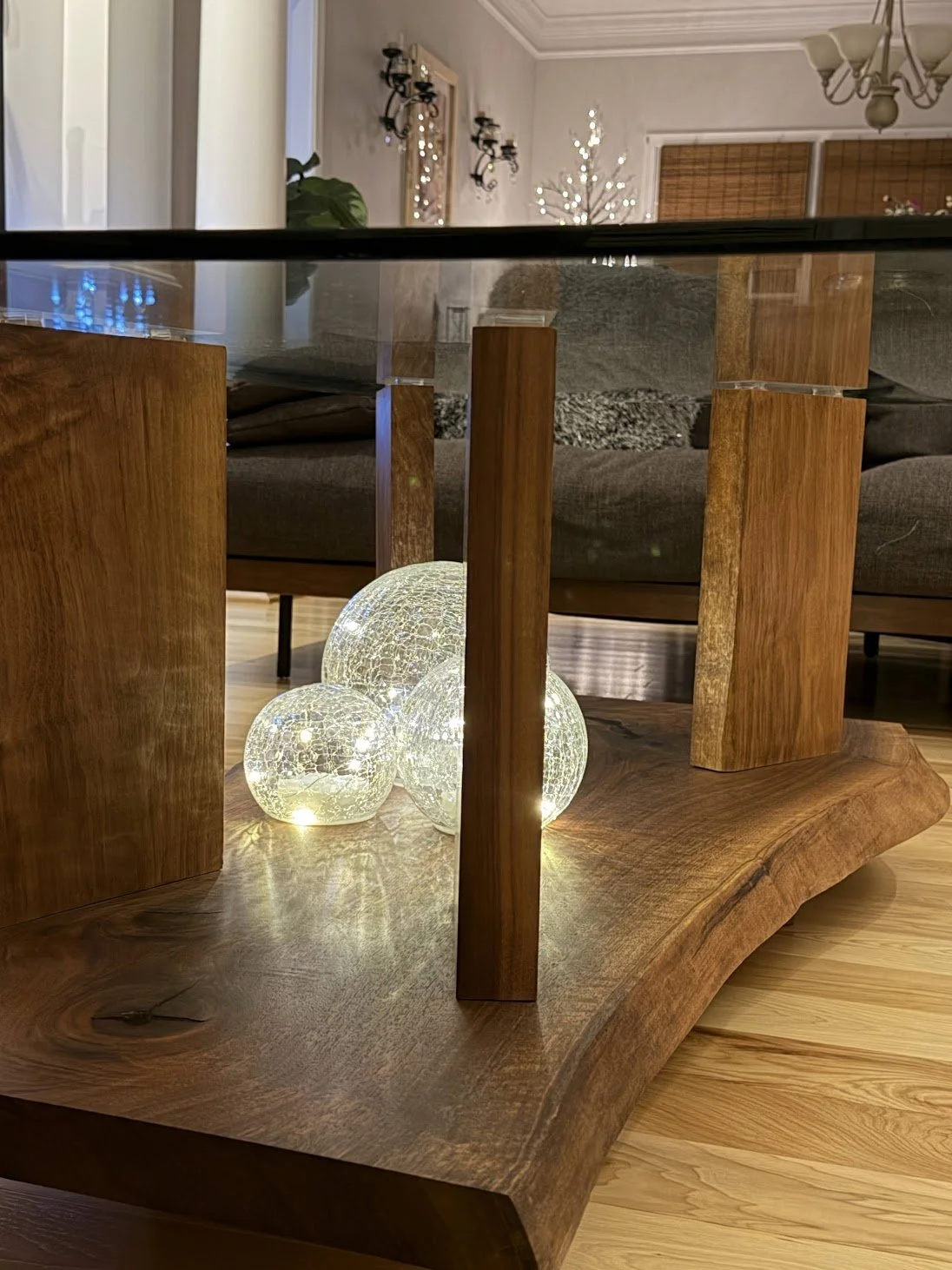 A close-up of a wooden table with a thick, natural edge, decorated with glass globe string lights underneath the table. In the background, there are chairs, a grey couch, a wall with decorative lighting fixtures, and a window.