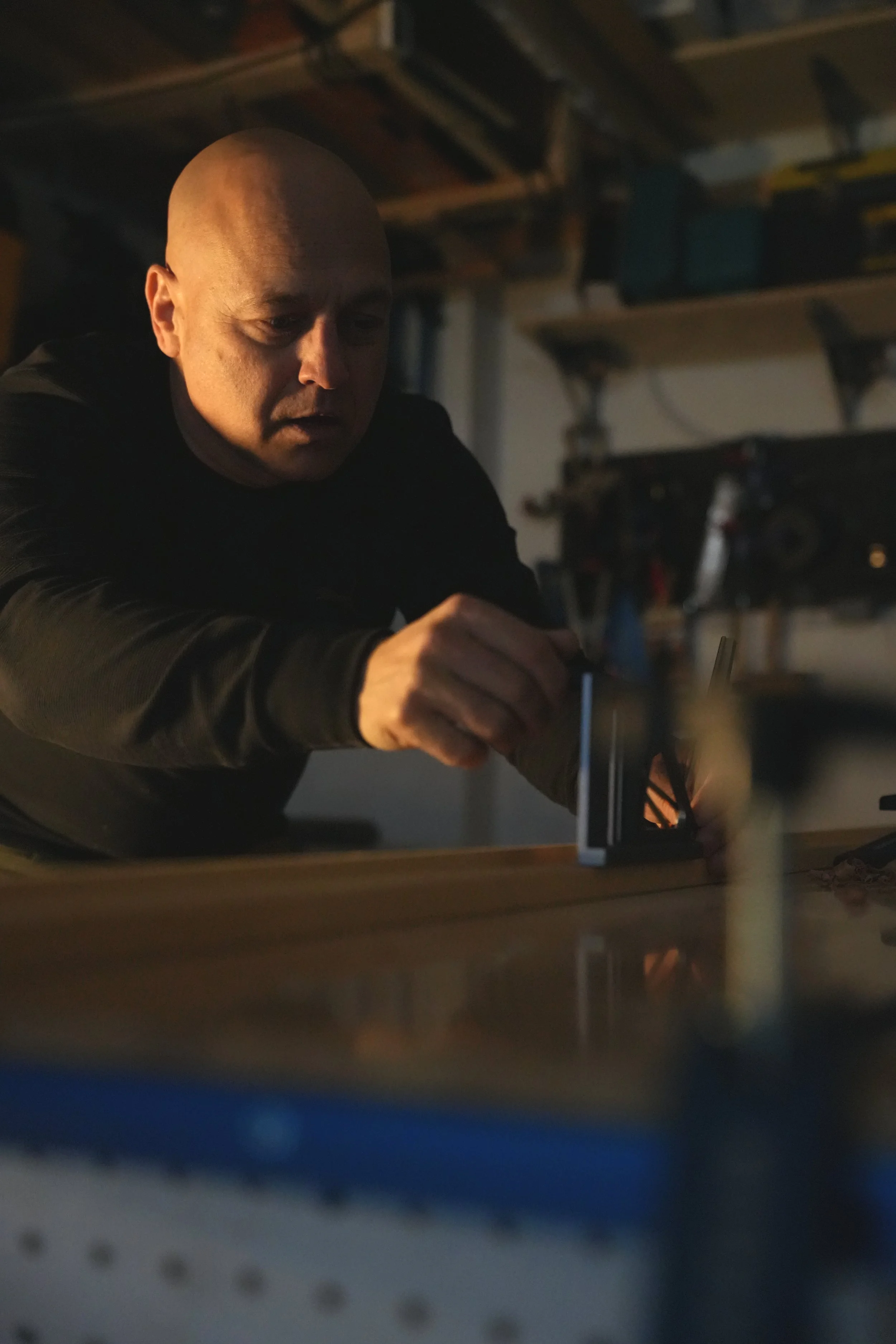 Bald man working in a garage or workshop, focusing on a task at a workbench, with equipment and shelves in the background.