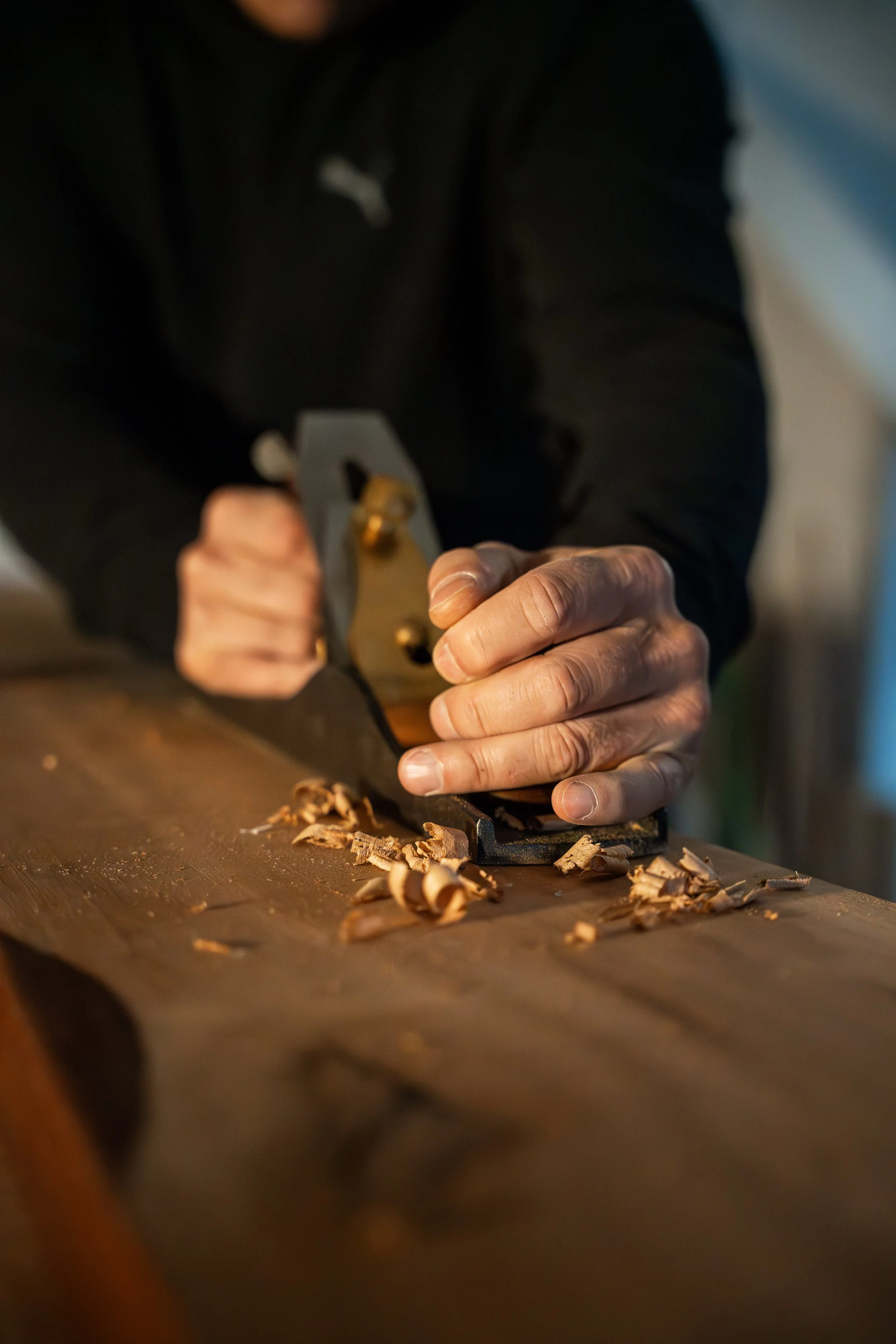 Close-up of a person using a hand plane on a wooden surface, shaving wood to make custom furniture from reclaimed wood