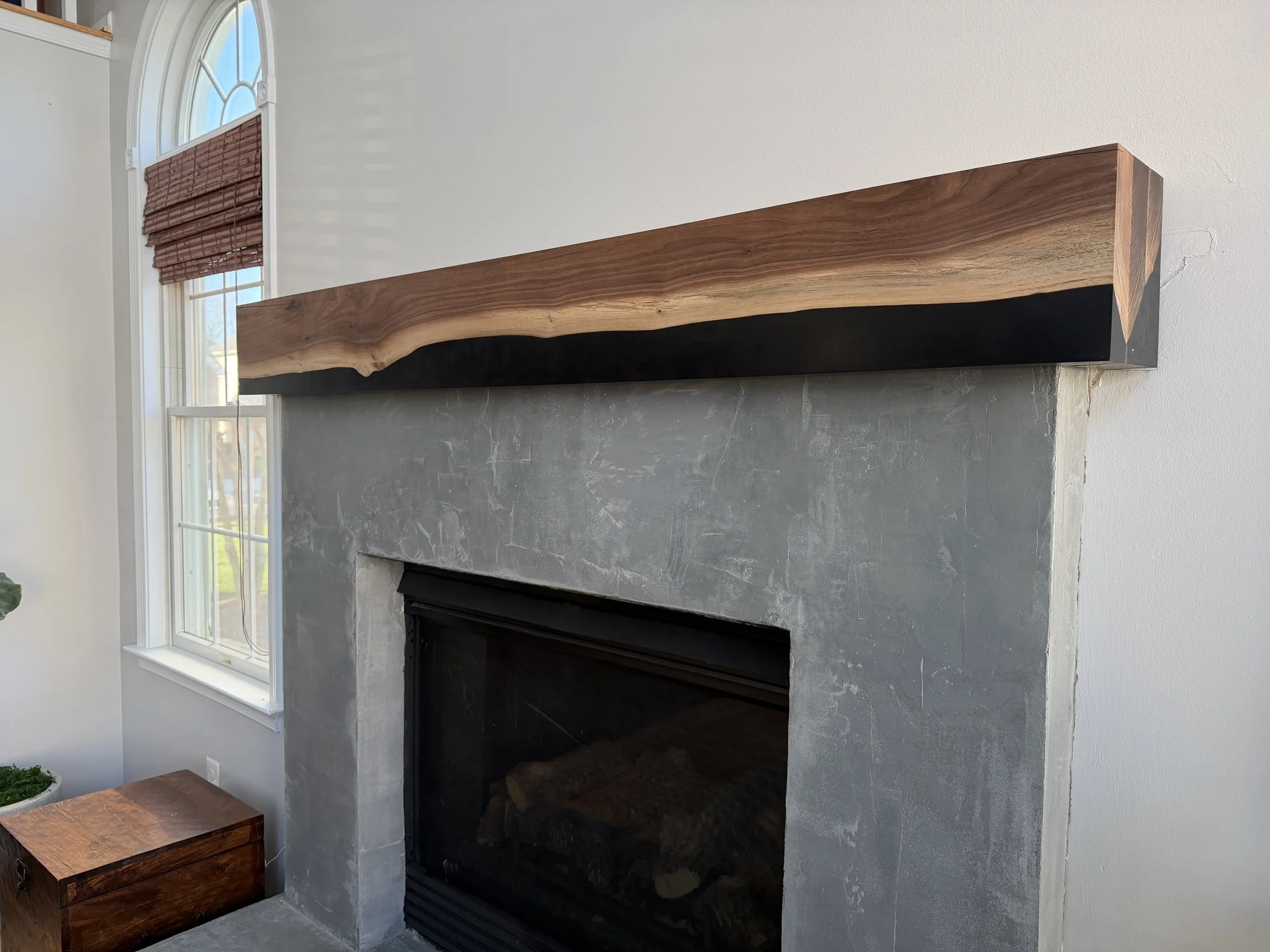 Close-up of a modern fireplace with a concrete surround and a wooden mantel above it in a room with large windows and white walls.