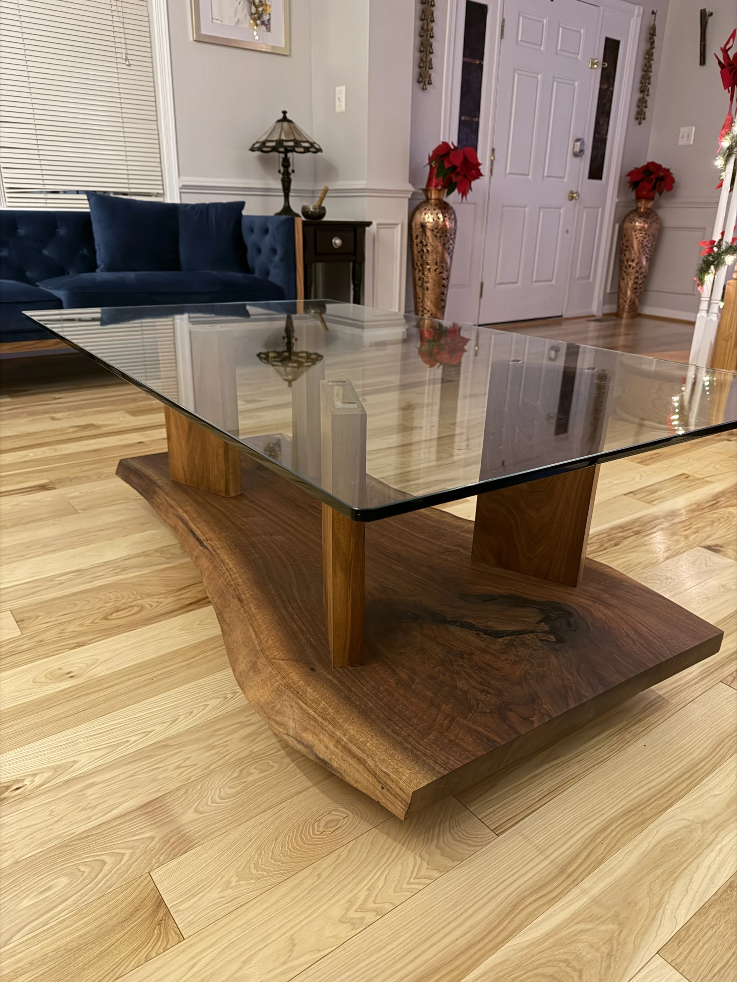 A modern coffee table with a glass top and wooden base in a living room with hardwood floors, a blue sofa, and decorative vases with poinsettias.