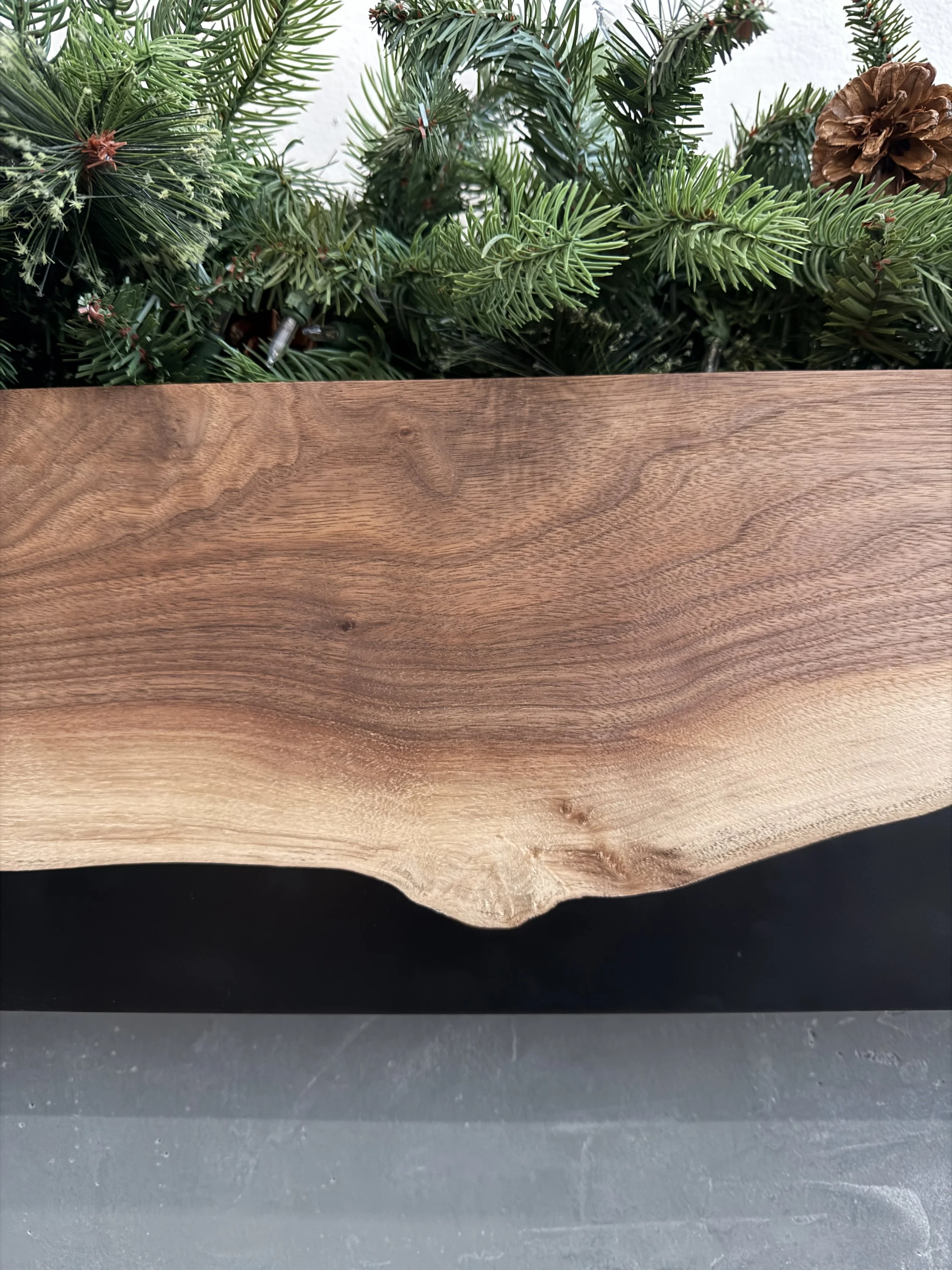 Close-up of a wooden tabletop with a natural edge, against a background of green pine garland with pinecones.