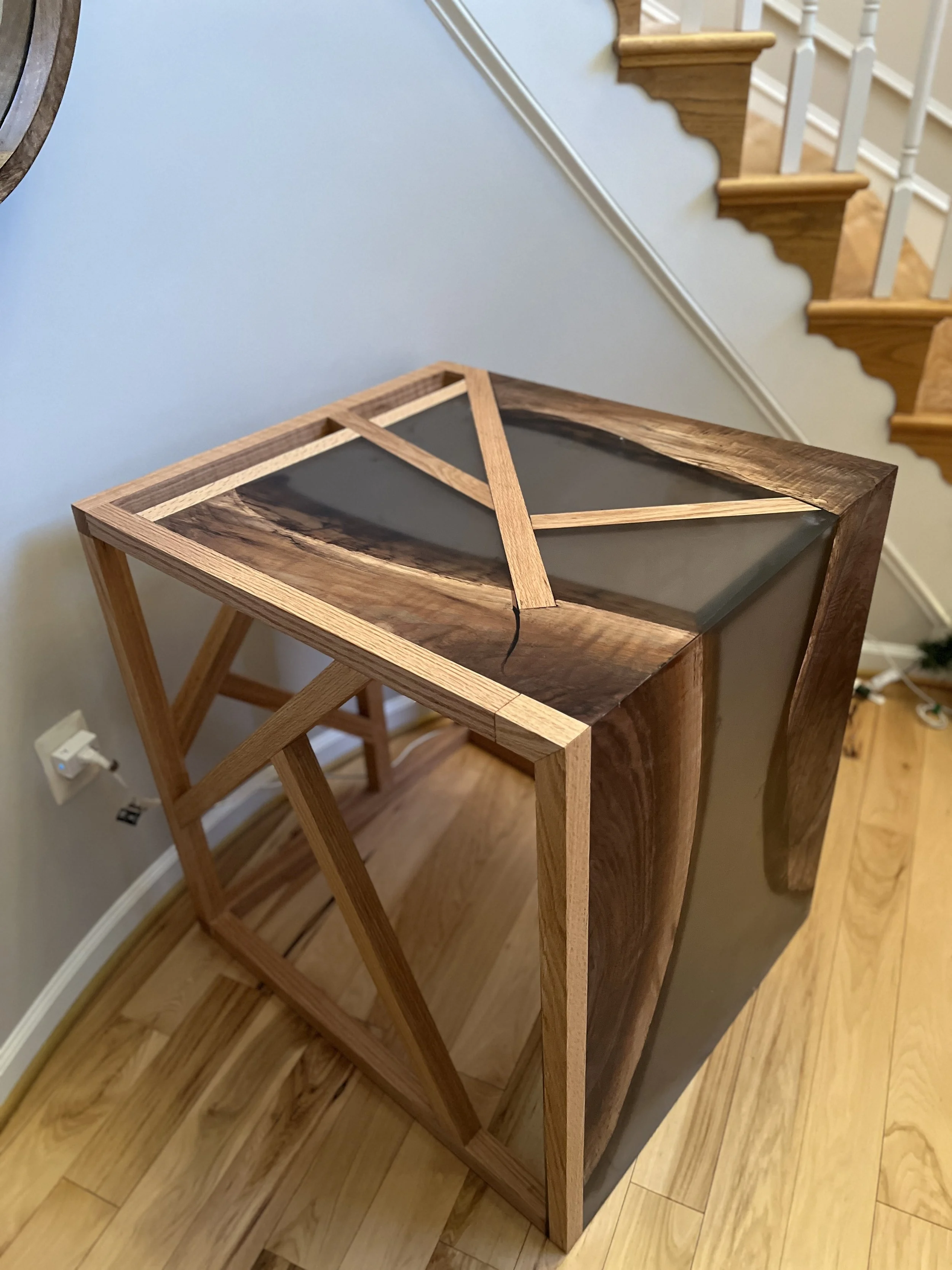 A wooden and resin table under construction, partially assembled, with a black resin top and wooden edges, placed in a room near a staircase.