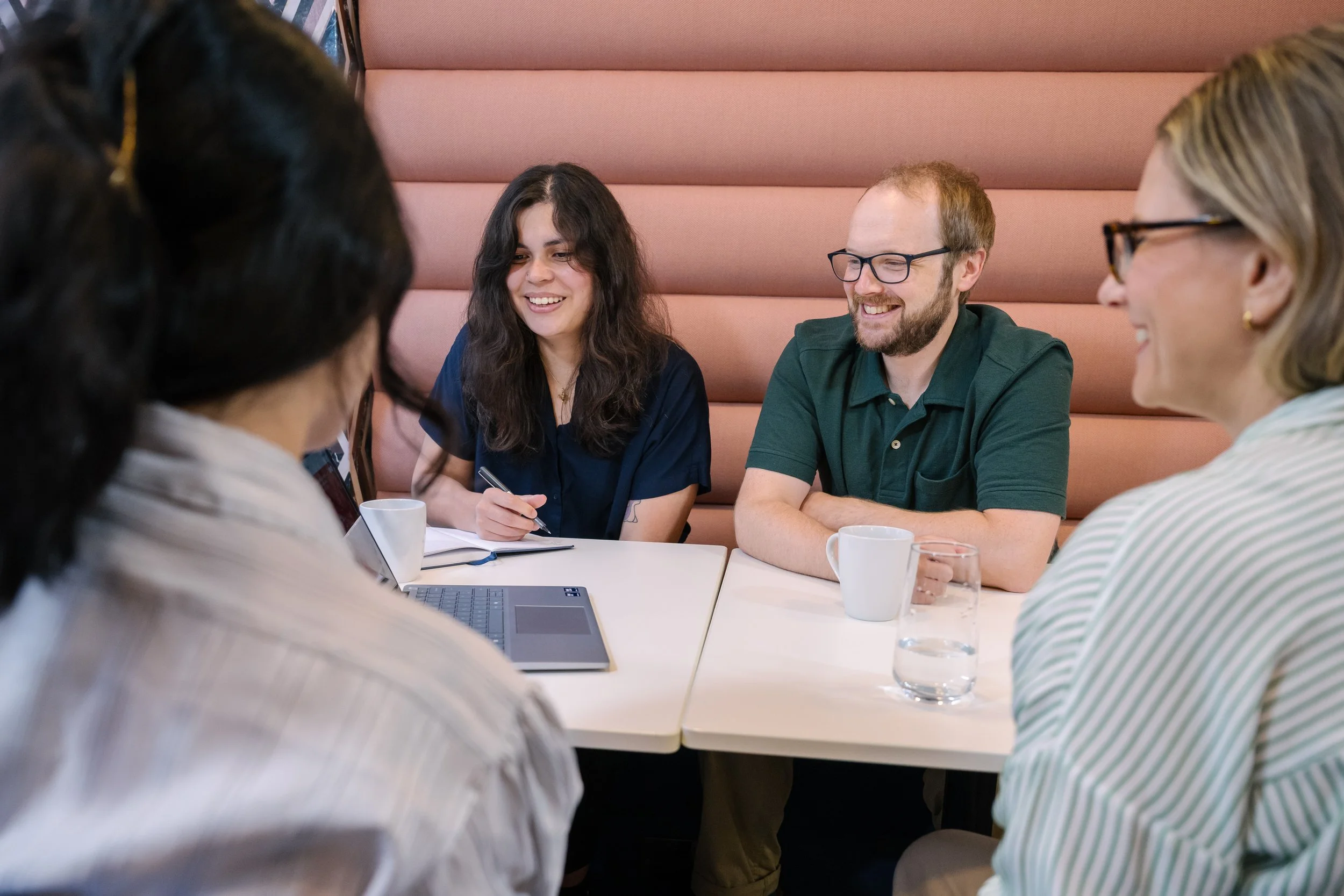 Group of four diverse people sitting around a table in a modern office or cafe, engaging in a discussion or meeting, with pink cushioned wall in the background.