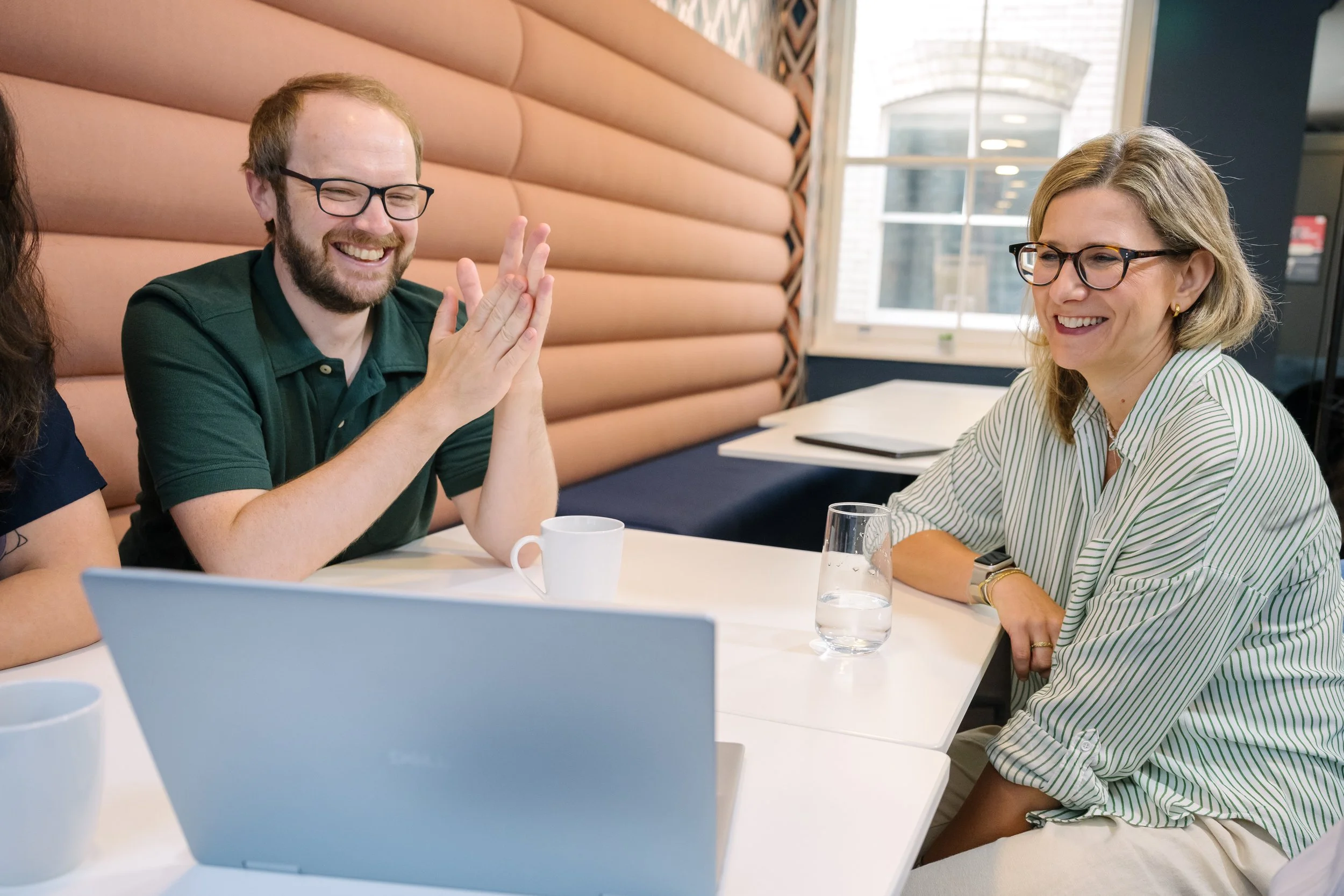 Two people sitting at a table, laughing and smiling, with a laptop and cups in front of them. The man has glasses, a beard, and is wearing a green shirt. The woman has glasses, blond hair, and is wearing a striped shirt.
