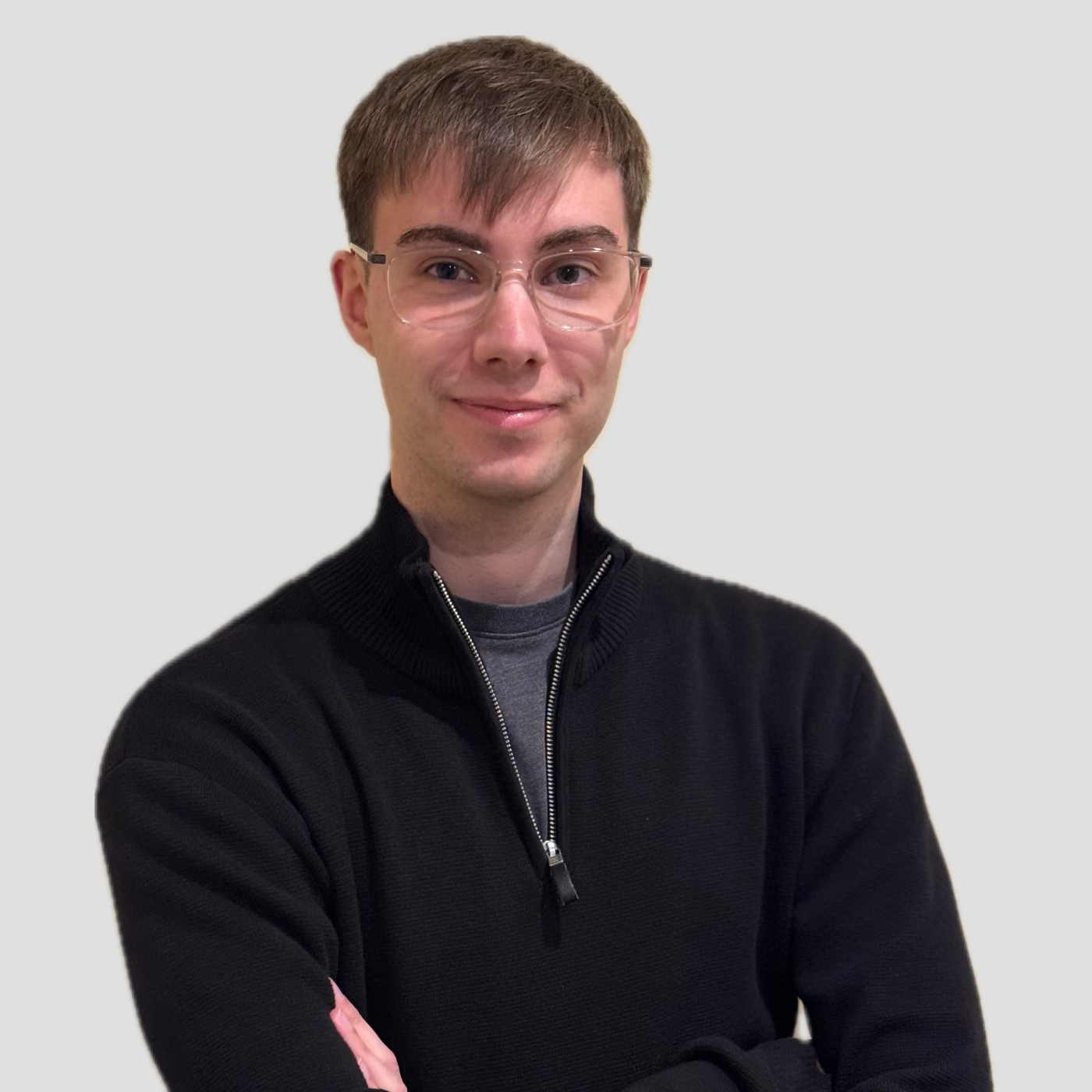 A young man with brown hair and glasses smiling, wearing a black zip-up sweater with a gray shirt underneath, standing with arms crossed against a plain light gray background.
