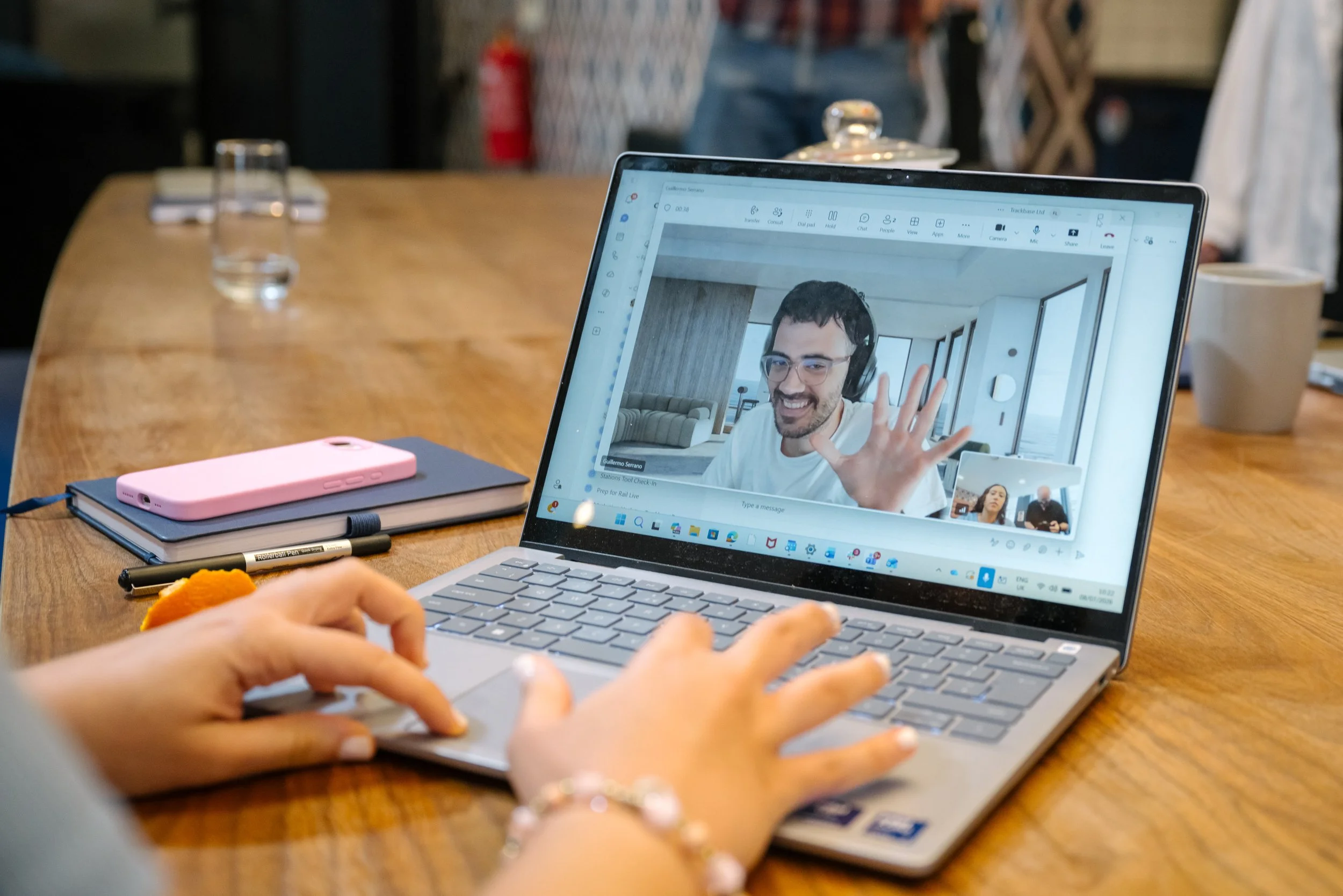 A person using a laptop in a meeting, on the screen is a video call with a man waving and smiling.