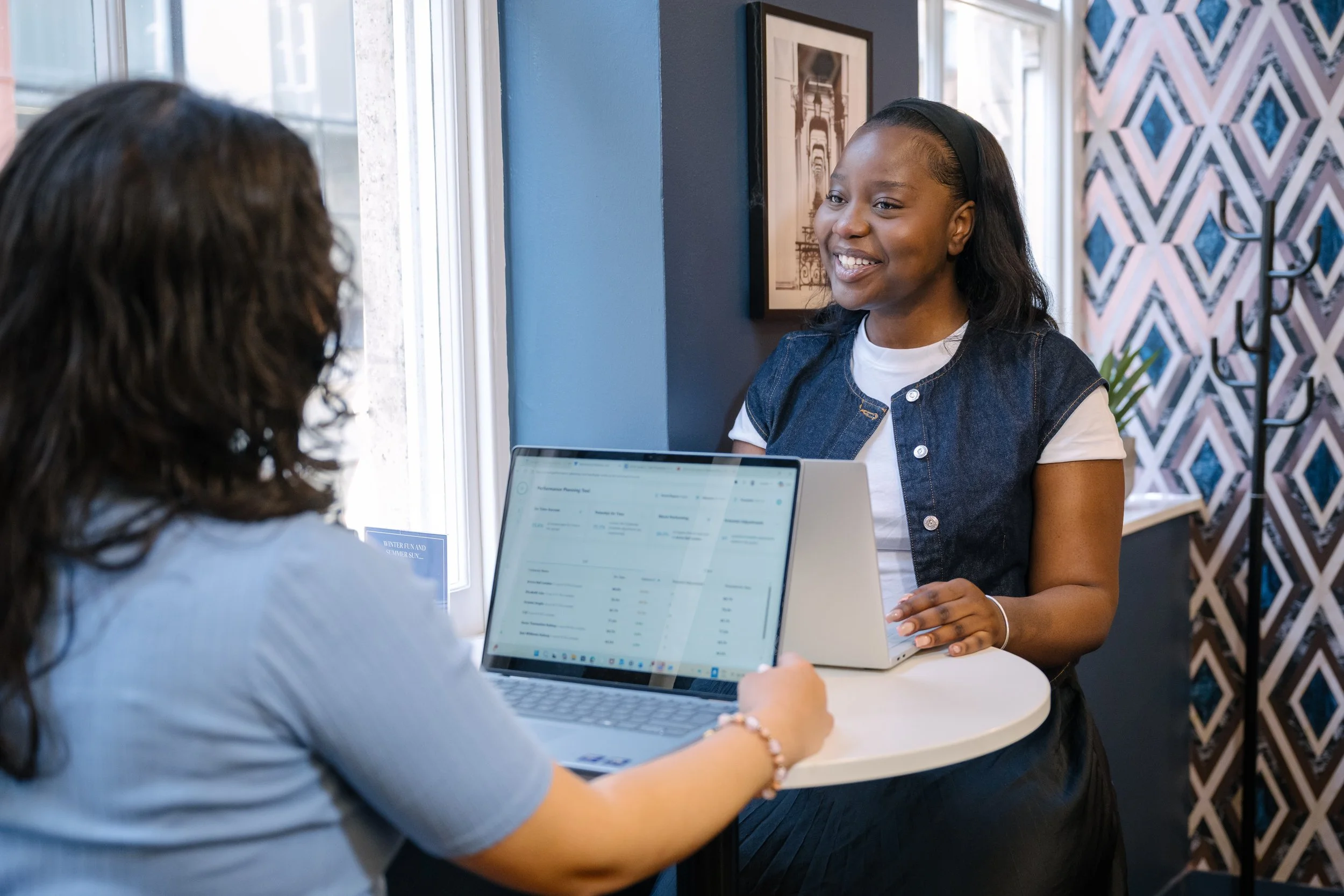 Two women having a conversation at a small round white table in a bright room, one woman smiling and the other showing her something on a laptop.