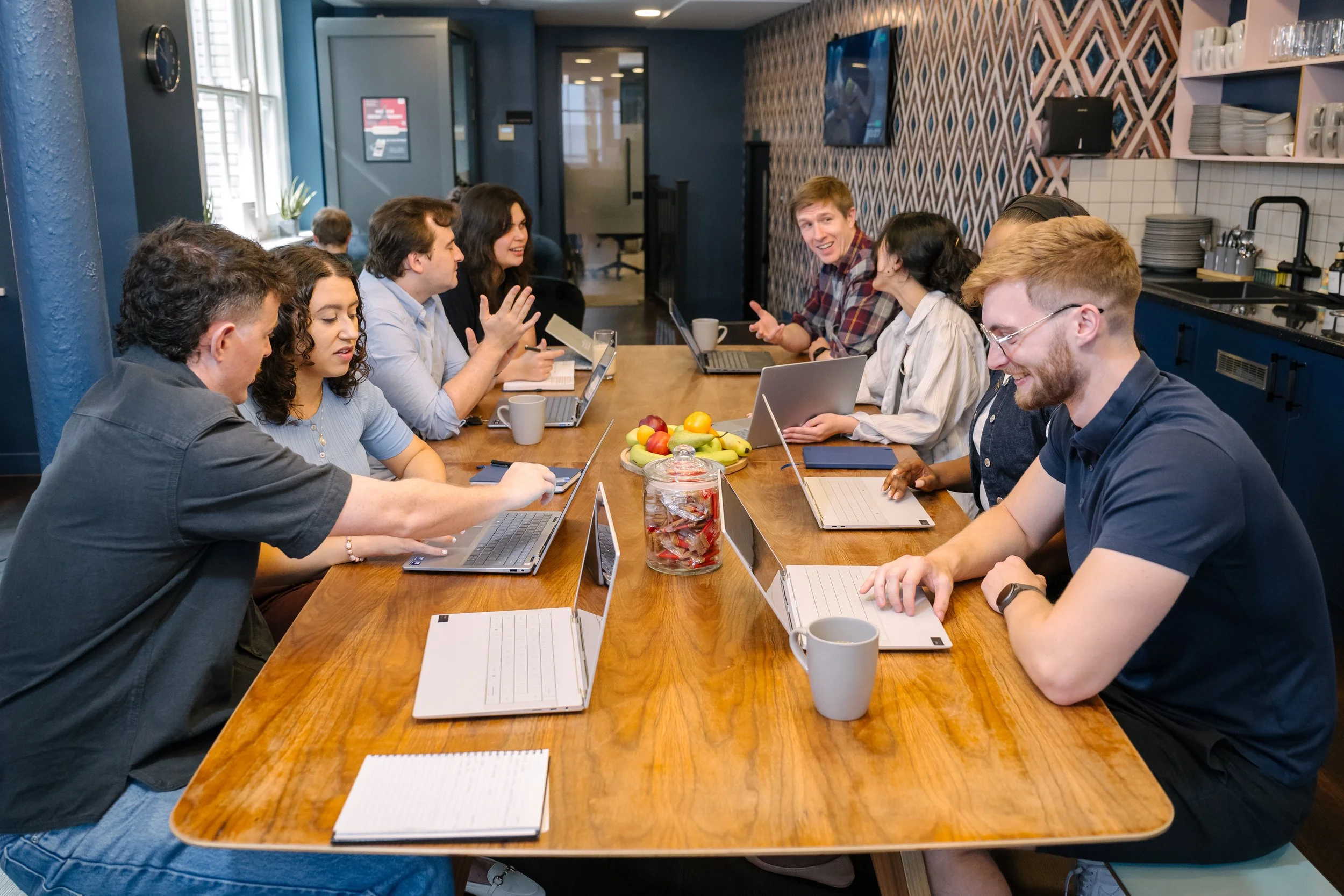 A group of ten people sitting around a long wooden table in a cozy, modern kitchen or dining area, working on laptops and engaging in conversation. There are notebooks, coffee mugs, and a jar of candies on the table, with a plate of fruit in the center.