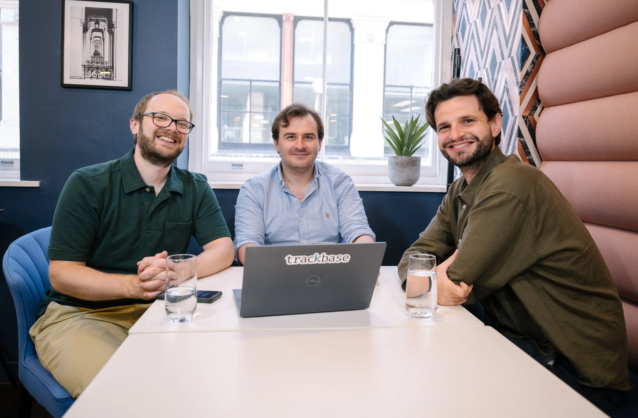 Three men sitting at a white table in a modern office space, smiling at the camera. They have glasses of water in front of them and a laptop displaying the Trackbase logo. The office has large windows, a potted plant, and colorful wall decor.