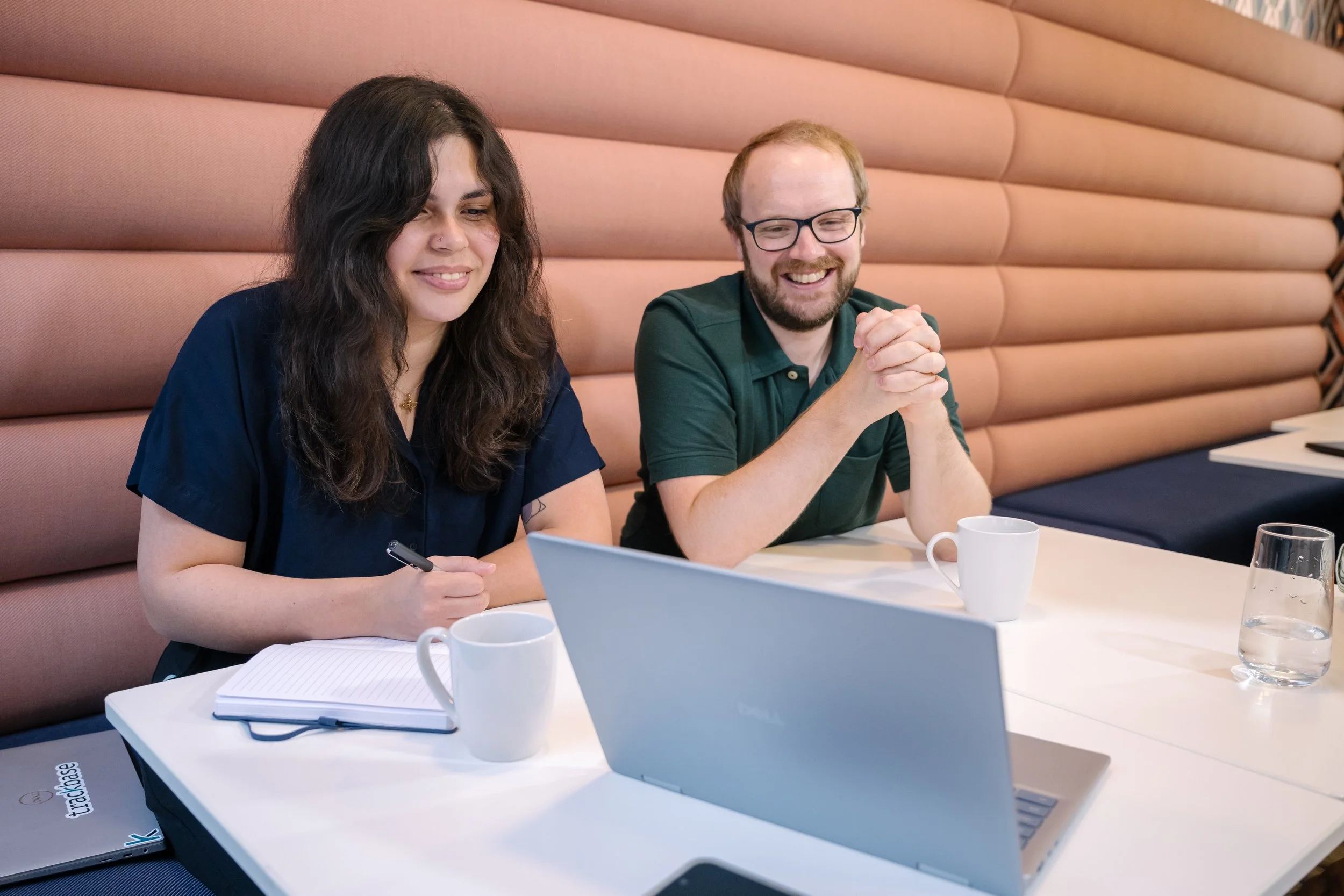 Two smiling people sitting at a table with a laptop, notebooks, and cups, in a room with pink padded wall panels.