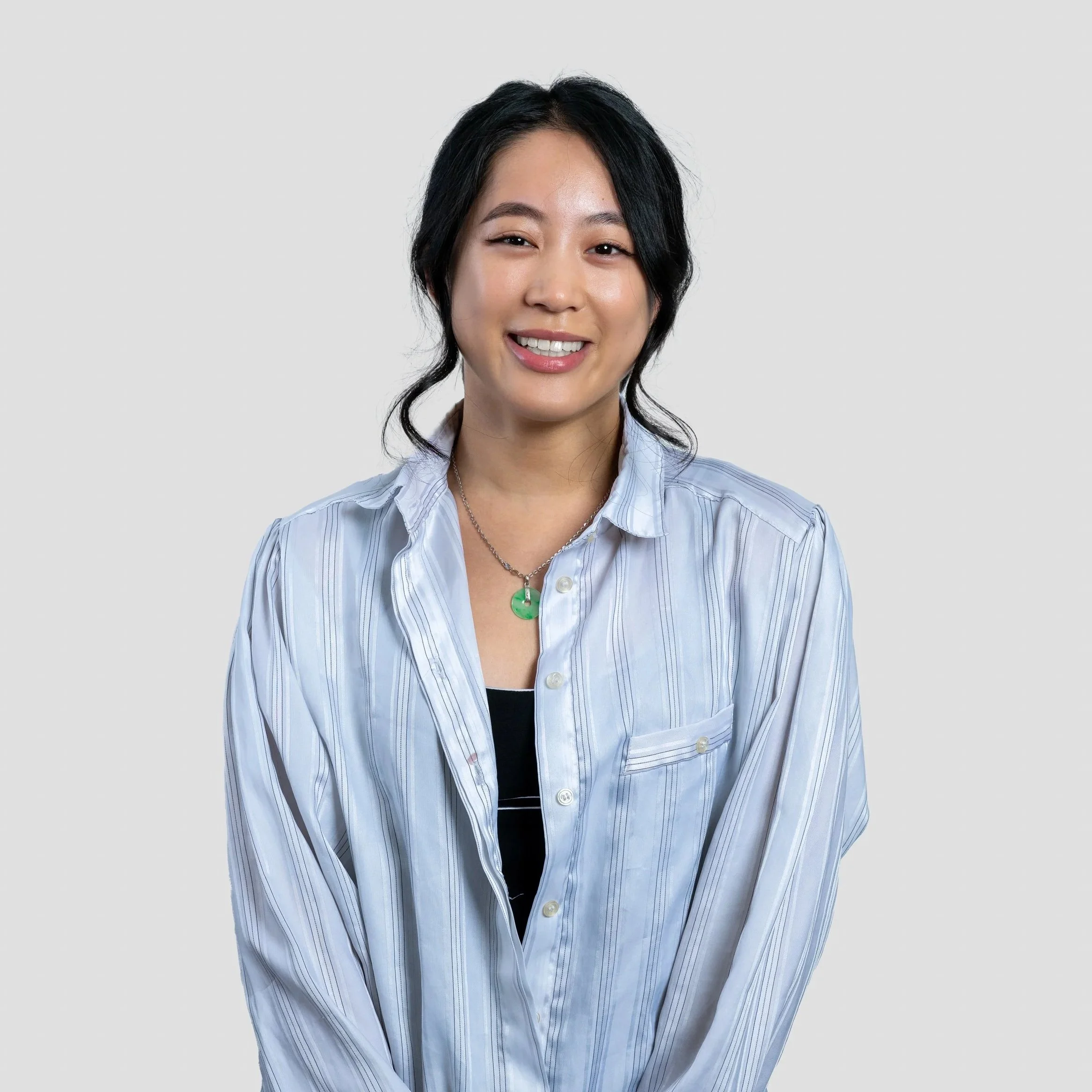 A woman with black hair styled in loose waves smiling at the camera, wearing a light blue striped shirt over a black top, with a green pendant necklace, against a plain white background.