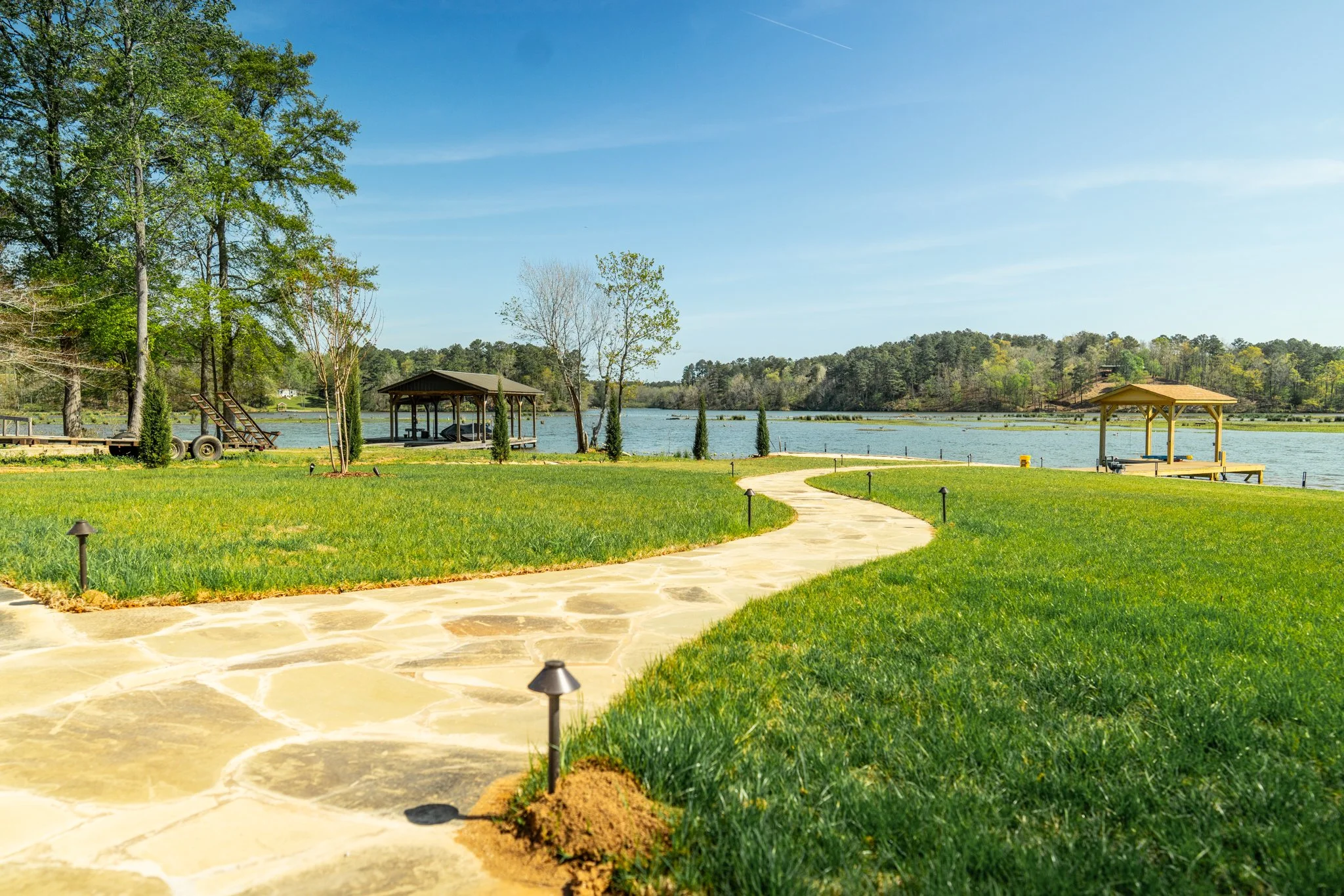 A scenic lakeside park with a stone path, green grass, trees, and several covered pavilions along the water, under a blue sky. Renovation in Birmingham, AL.