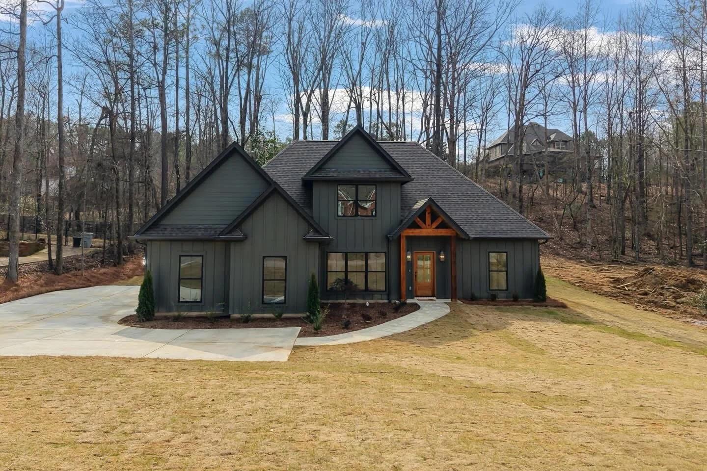 A modern two-story house with dark gray siding, a gabled roof, and a small front porch surrounded by leafless trees. The house has a curved walkway leading to the front door and a concrete driveway on the left. In the background, there are more houses on a wooded hill.