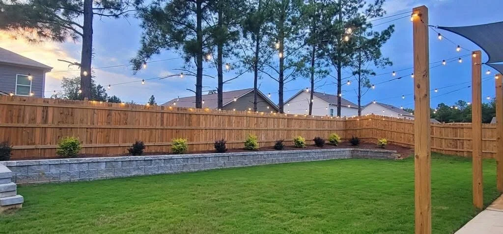 A backyard with a fresh wooden fence, green lawn, decorative string lights, and small bushes along a stone garden bed during dusk in Birmingham, AL, and Shelby, AL.