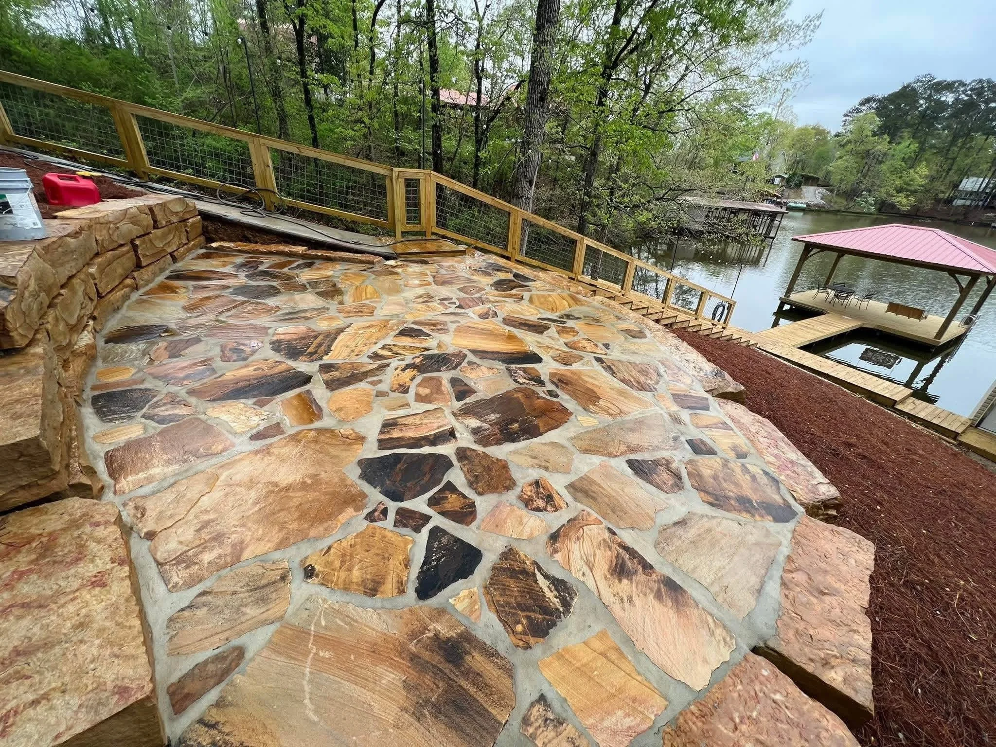 A stone patio with a wooden railing and steps leading down to a dock on a river. There is a gazebo with a red roof over the dock. Trees surround the area, and there are boats visible on the river. Outdoor remodel in Birmingham, Alabama