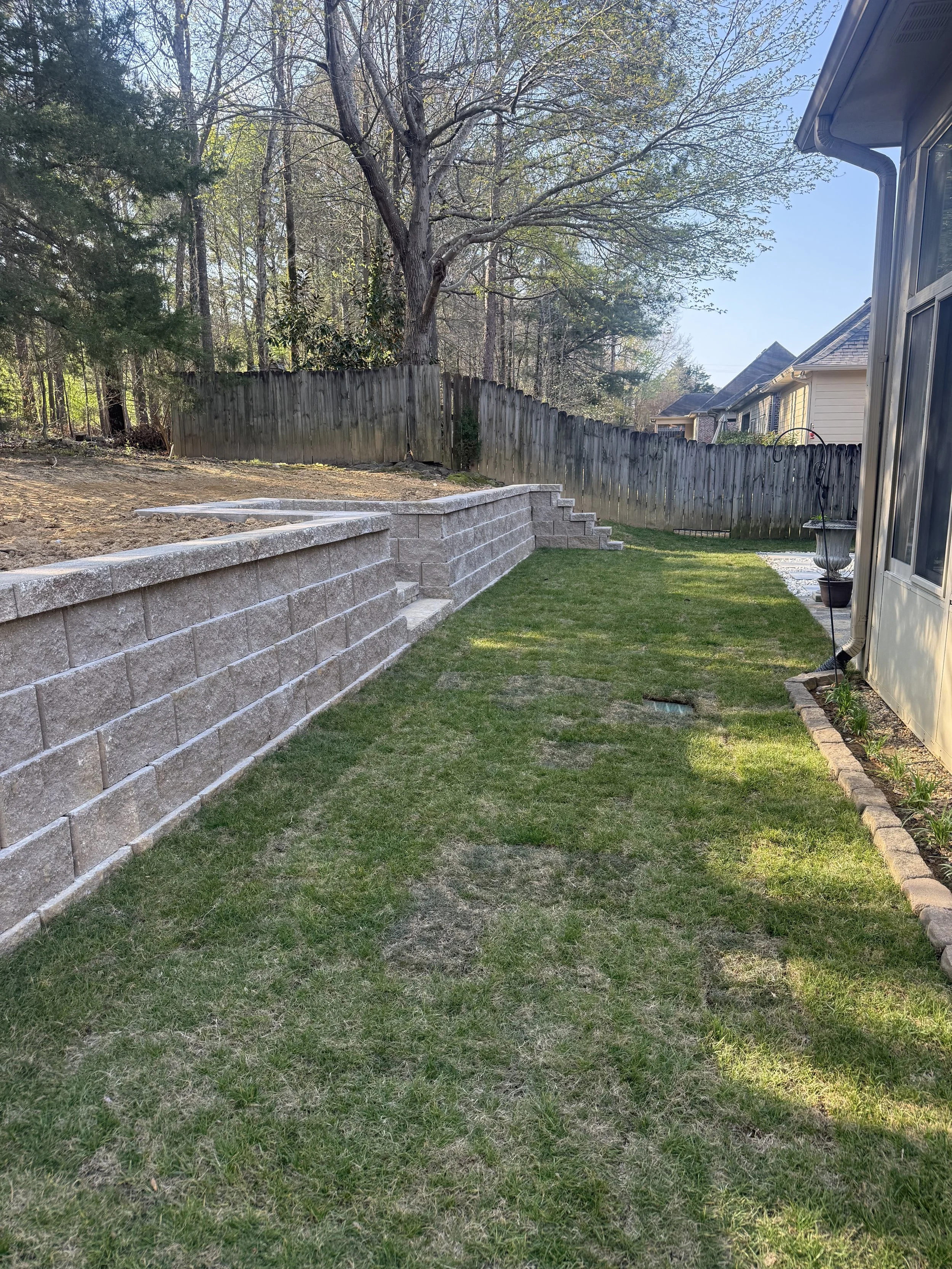 Backyard garden with a stone retaining wall, green grass, a large tree, and a wooden fence, with neighboring house visible in the background in Birmingham, AL.
