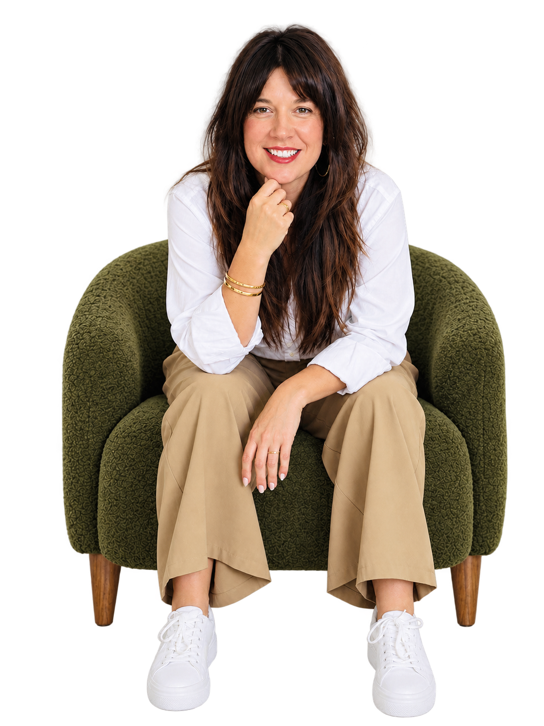Rebecca Tye with long brown wavy hair, wearing a white shirt, beige pants, and white sneakers, sitting on a green textured armchair, smiling with her chin resting on her hand.