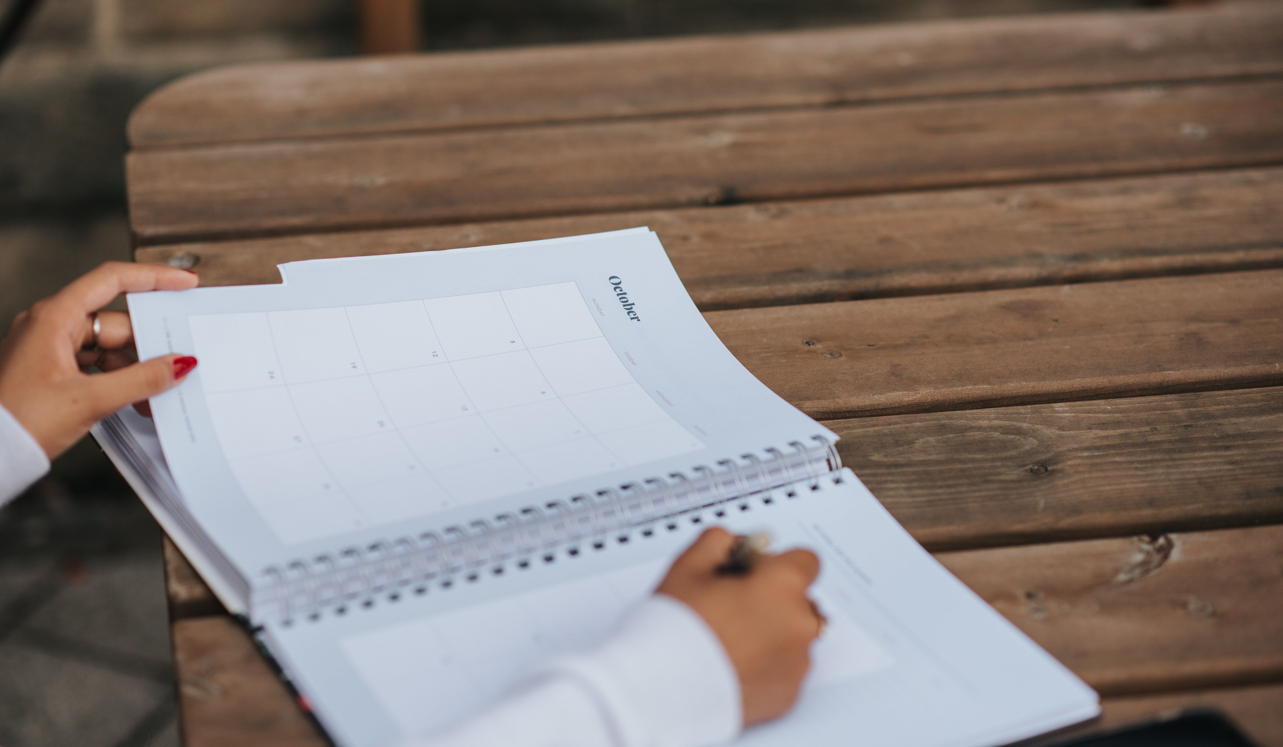 A person's hand with red nail polish is holding an open spiral notebook with a calendar page titled "October" on a wooden picnic table.