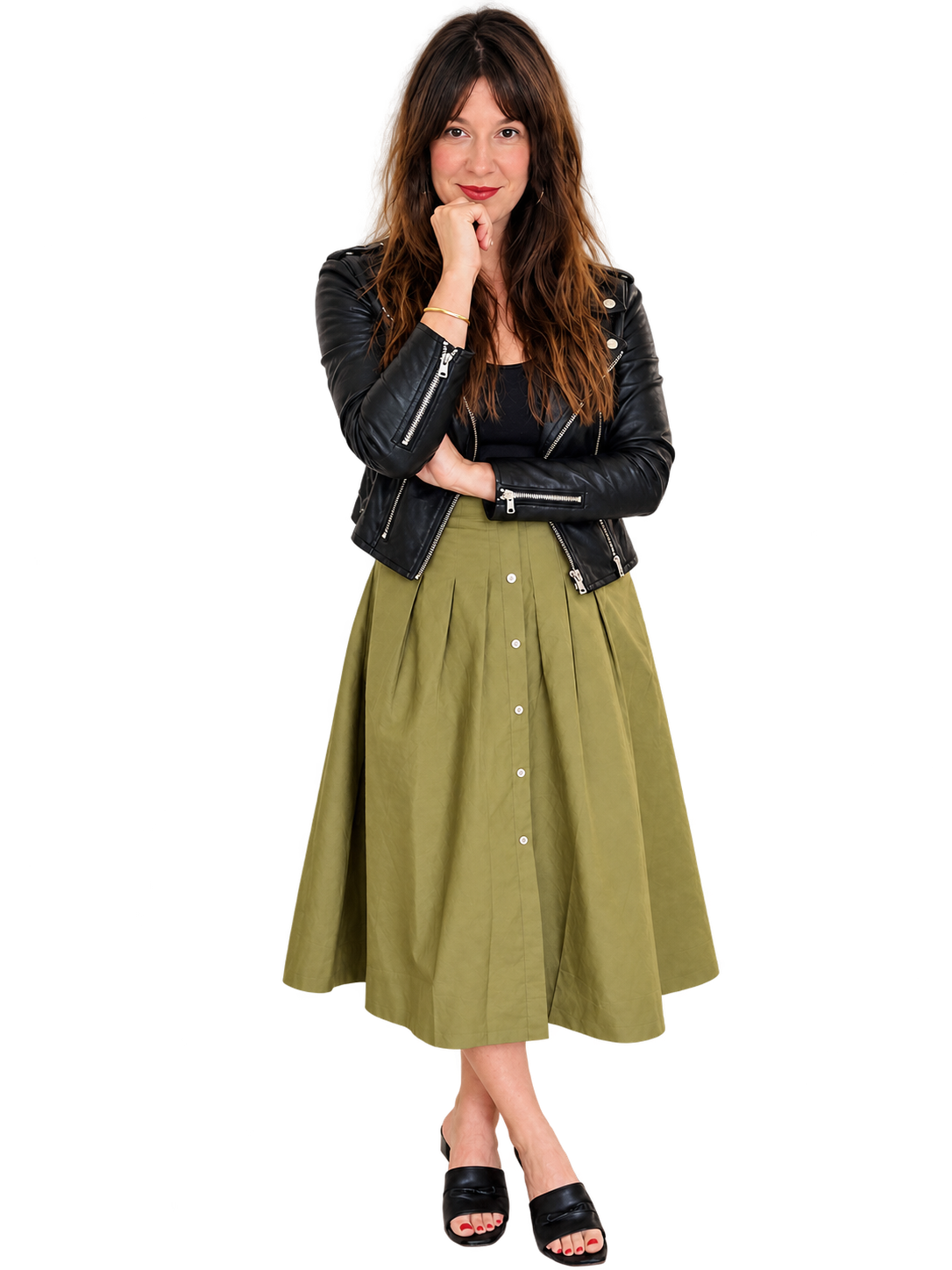 Rebecca Tye with long brown hair wearing a black leather jacket, black top, green pleated skirt with buttons, and black sandals, standing with her hand on her chin.
