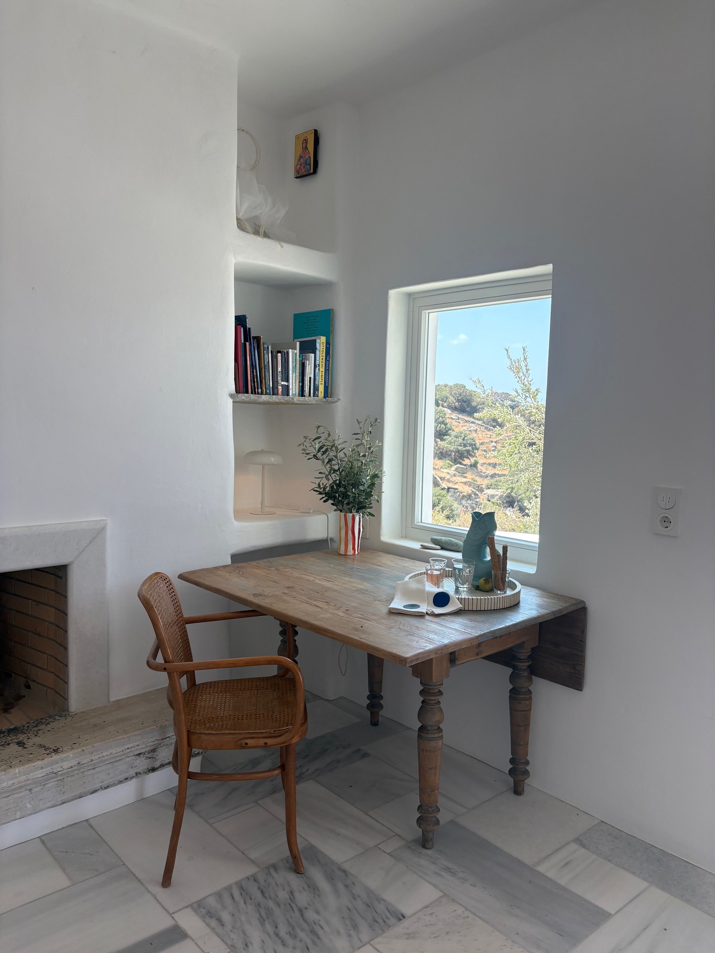 Interior of a cozy, minimalist room with white walls and a marble floor, featuring a wooden table by a window with a small plant, a tray, and a few glasses, and a wicker chair.
