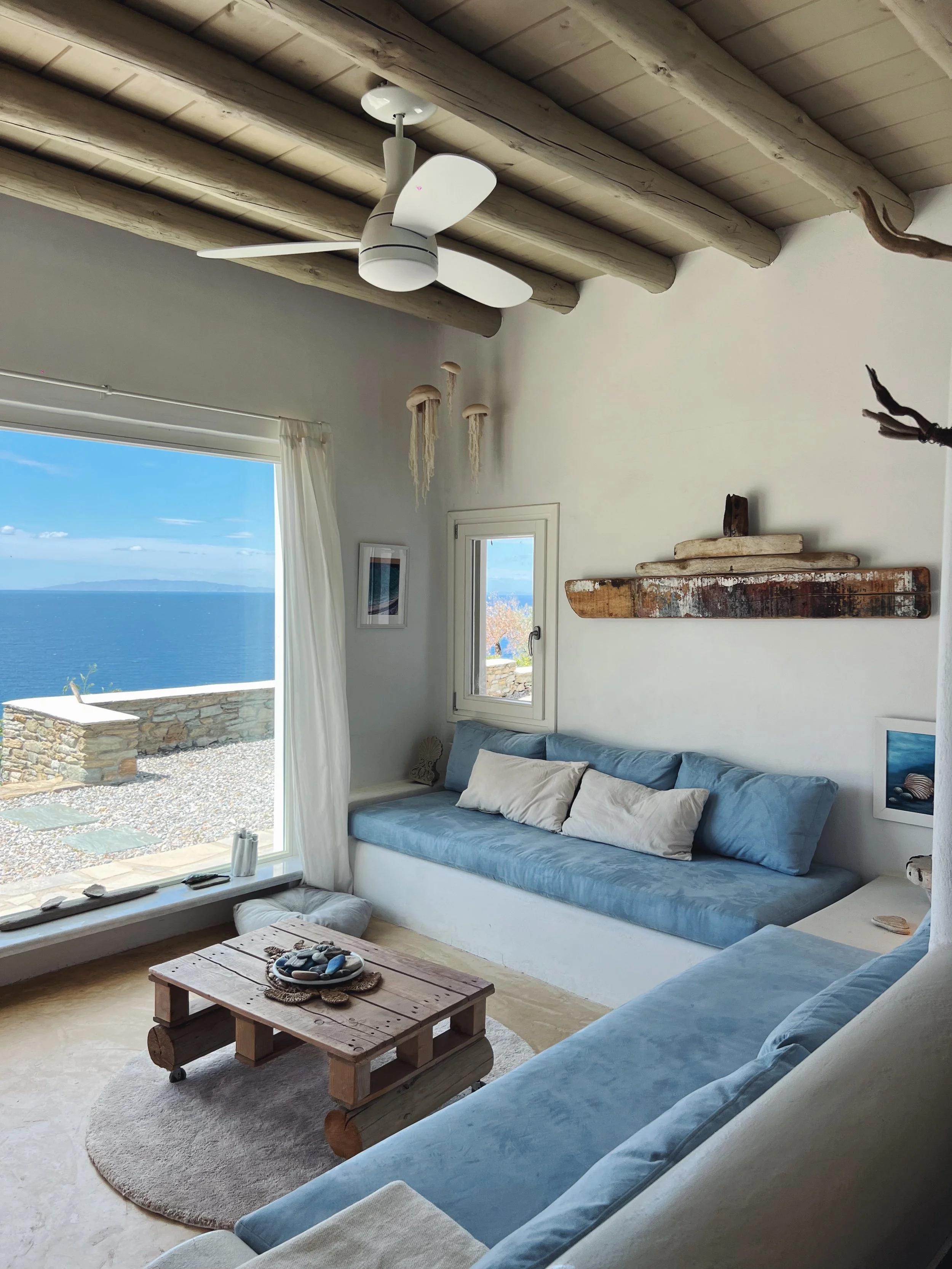 Cozy living room with sea view, featuring white walls, wooden ceiling beams, and light blue cushioned seating. A small window and a large glass door with white curtains open to an outdoor patio with a stone wall and ocean view. Decor includes hanging sea-themed art, a rustic wooden shelf, and a coffee table made from a pallet with stones on it. A white ceiling fan hangs from the ceiling.