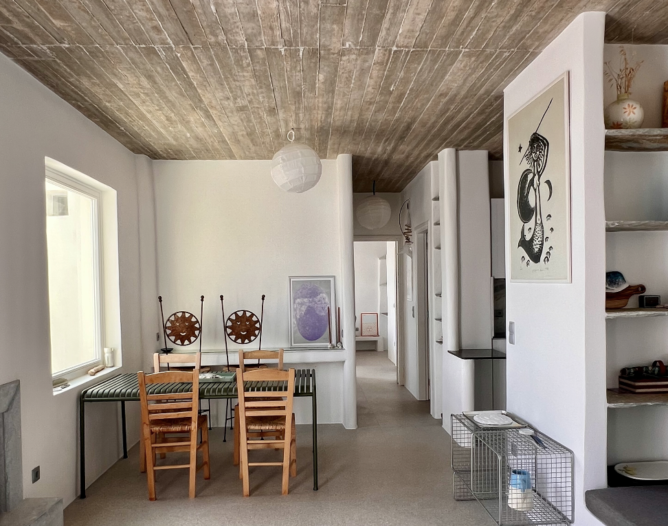 Interior of a modern home featuring a dining area with a table and four wooden chairs, a white wall with framed artwork, built-in shelves with decorative items, and a hallway with doors leading to other rooms.