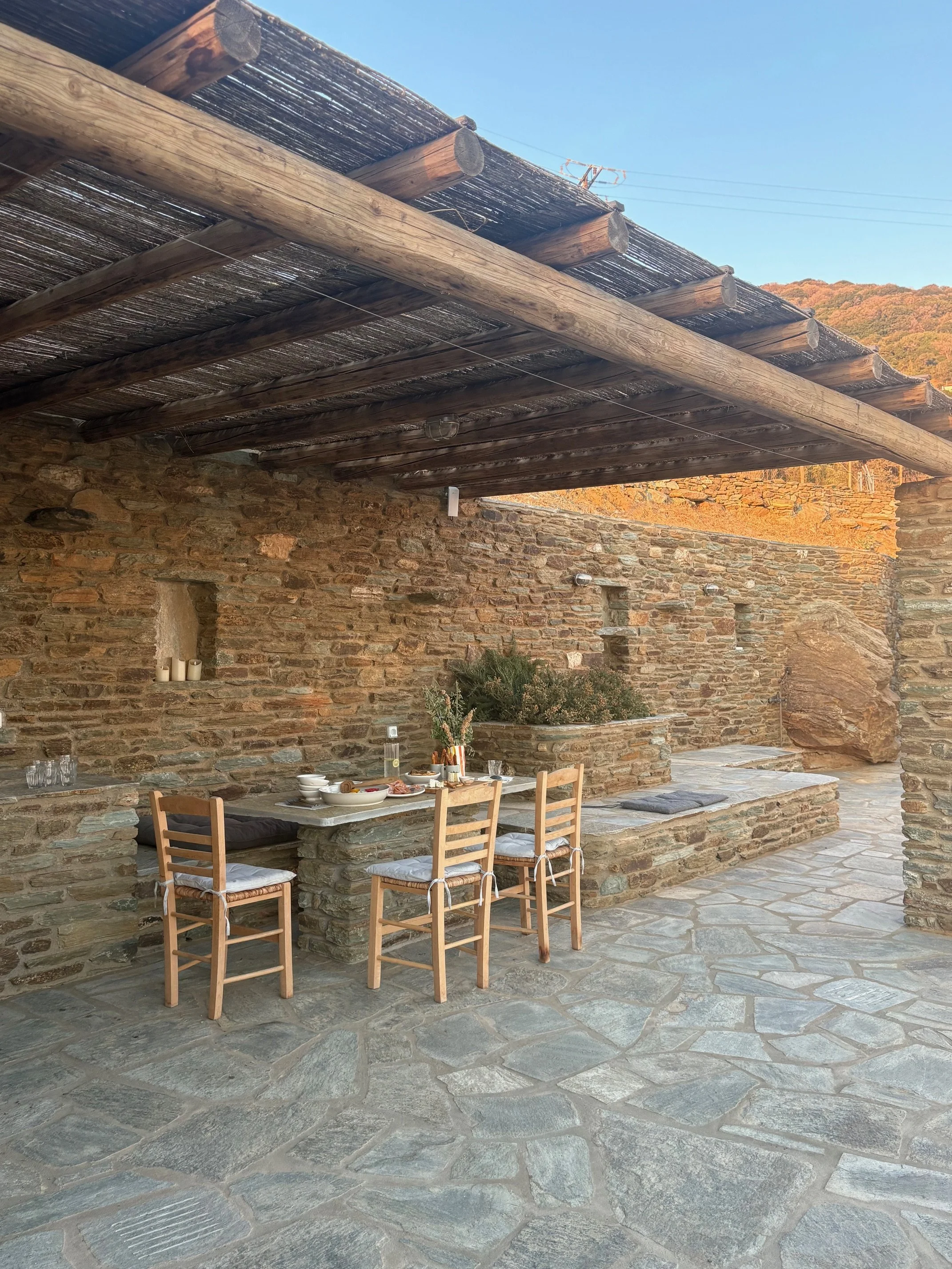 An outdoor dining area with a stone table and four wooden chairs under a wooden pergola. The table has dishes and utensils, and the area is surrounded by stone walls and a stone floor, with a scenic background of mountains and blue sky.