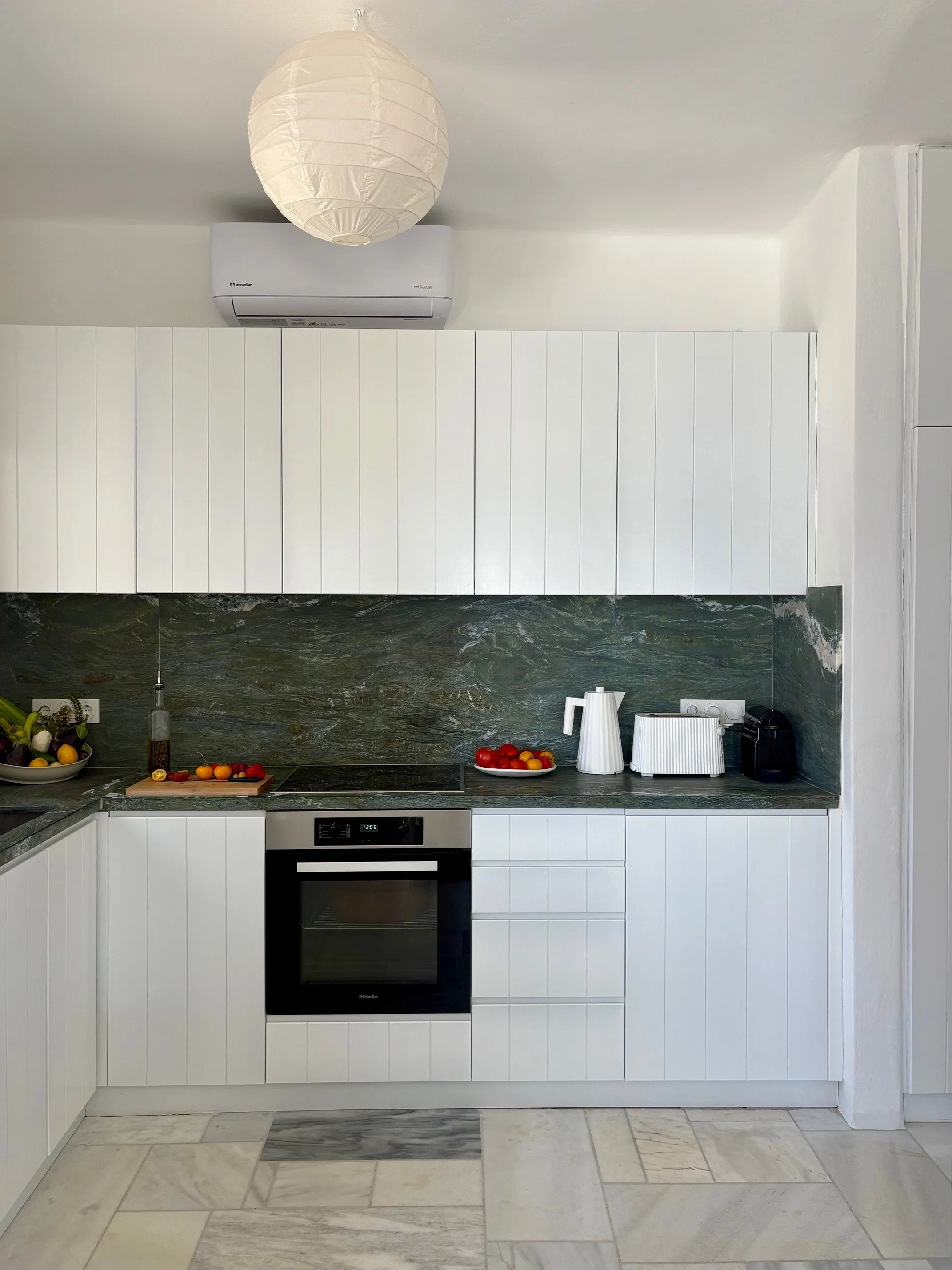 Modern kitchen with white cabinets, dark green marble backsplash, countertop with tomatoes, a white pitcher, toaster, and coffee machine. Overhead paper lantern light and wall-mounted air conditioner.