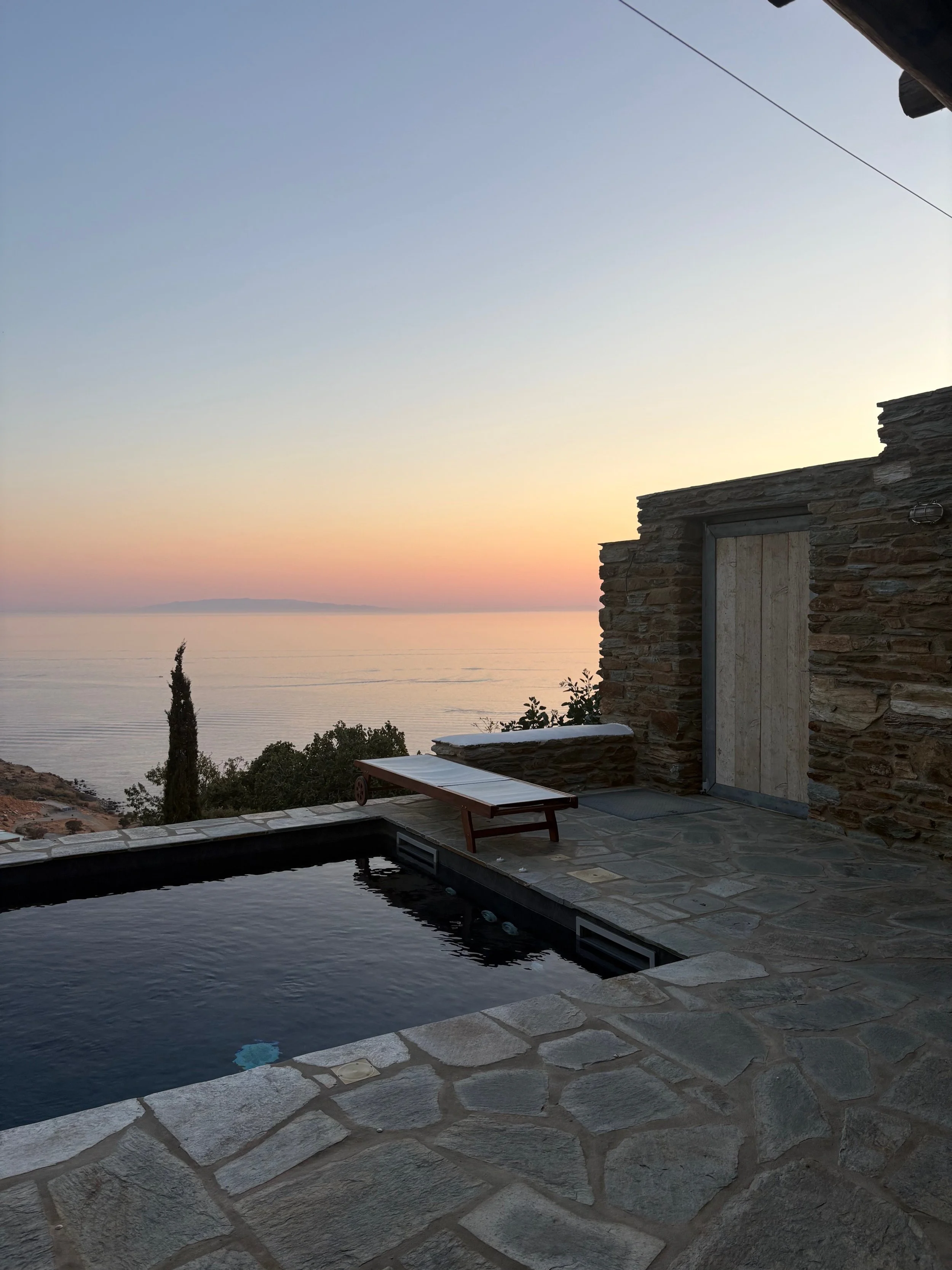 Sunset view over the ocean from a stone patio with a small pool, a sun lounger, and a stone building with a closed wooden door.