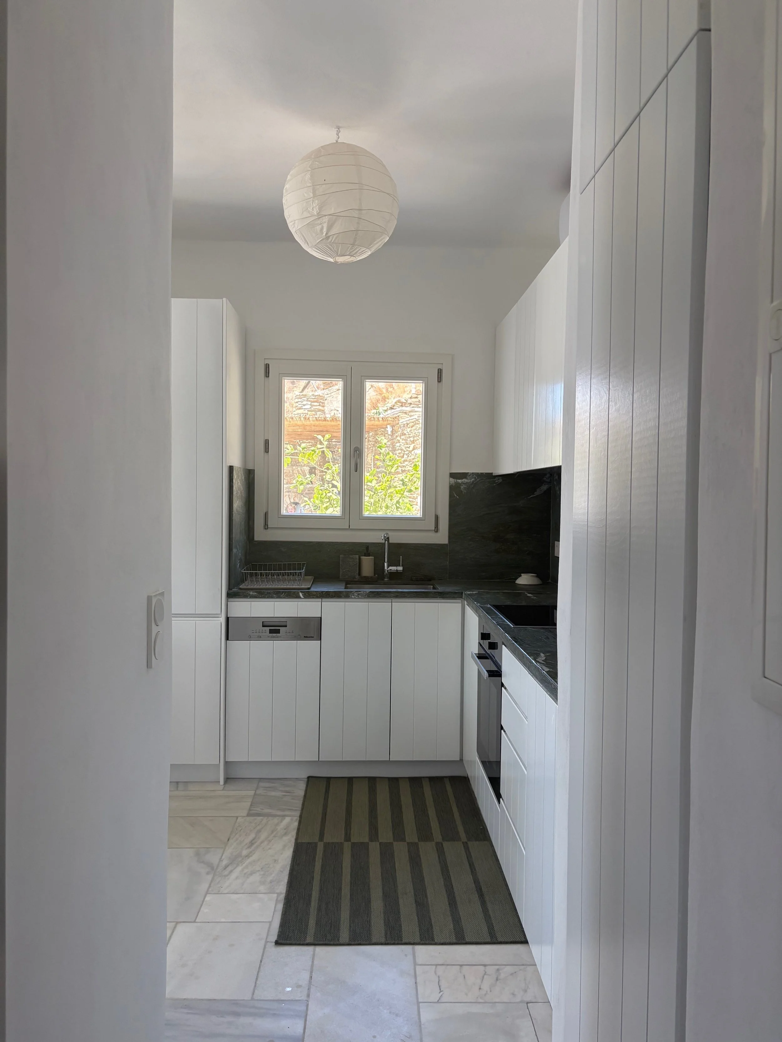 A small, modern kitchen with white cabinets, gray marble countertops, and a window overlooking greenery. A black and green striped rug lies on the tile floor, with a paper lantern hanging from the ceiling.