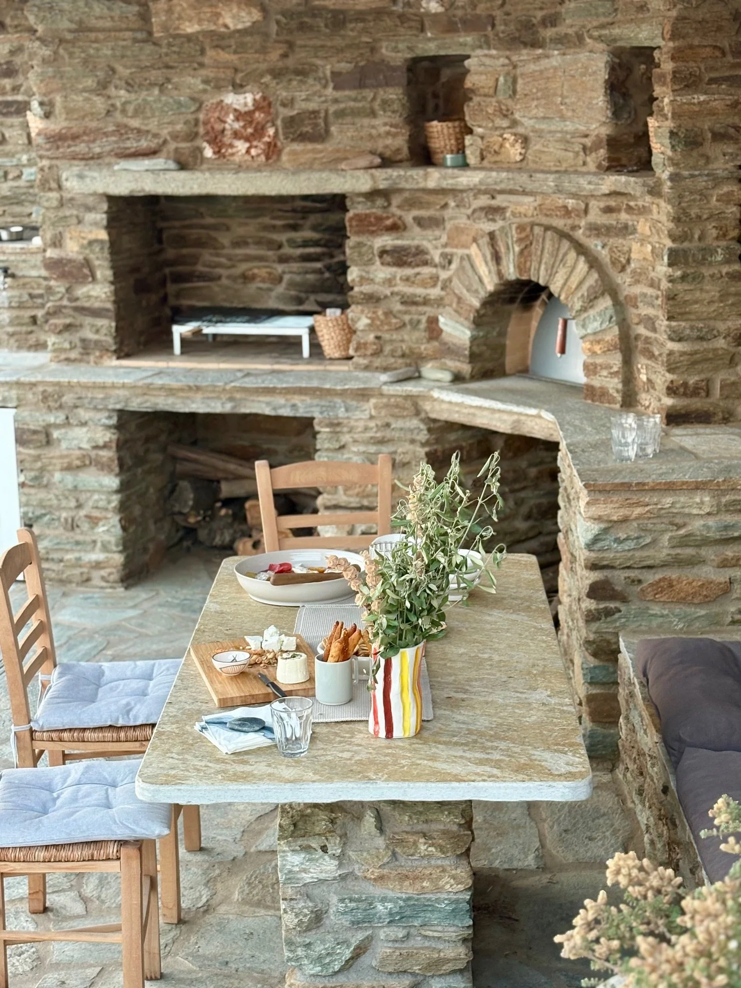A rustic stone indoor dining area with a stone table set with a large salad, small bowls, a wooden cutting board, and a striped vase with greenery, surrounded by wooden chairs with cushions and a built-in stone fireplace in the background.