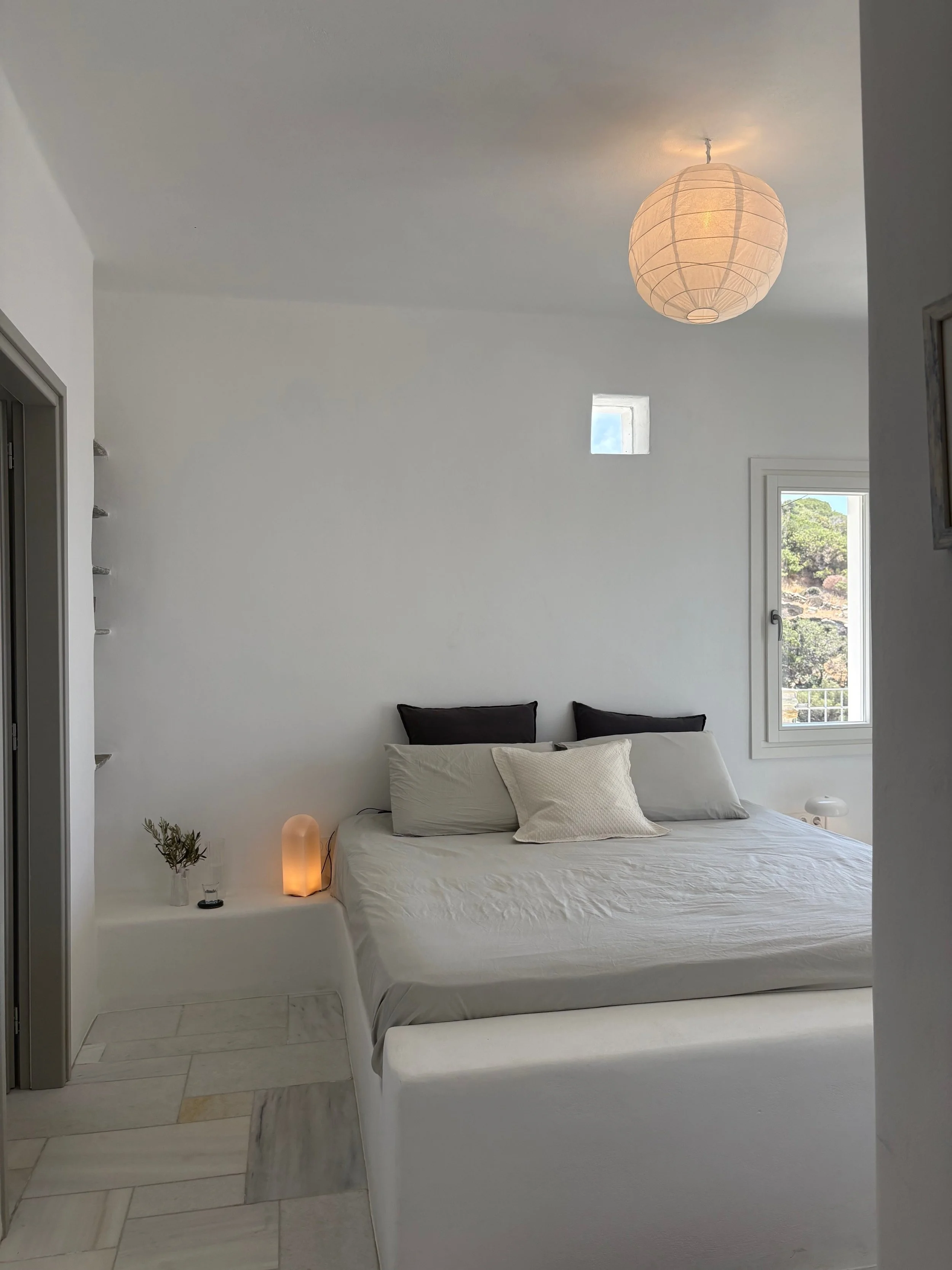 Minimalist bedroom with a white bed, black and white pillows, a small side table with a lamp and plant, window with view, and soft lighting.